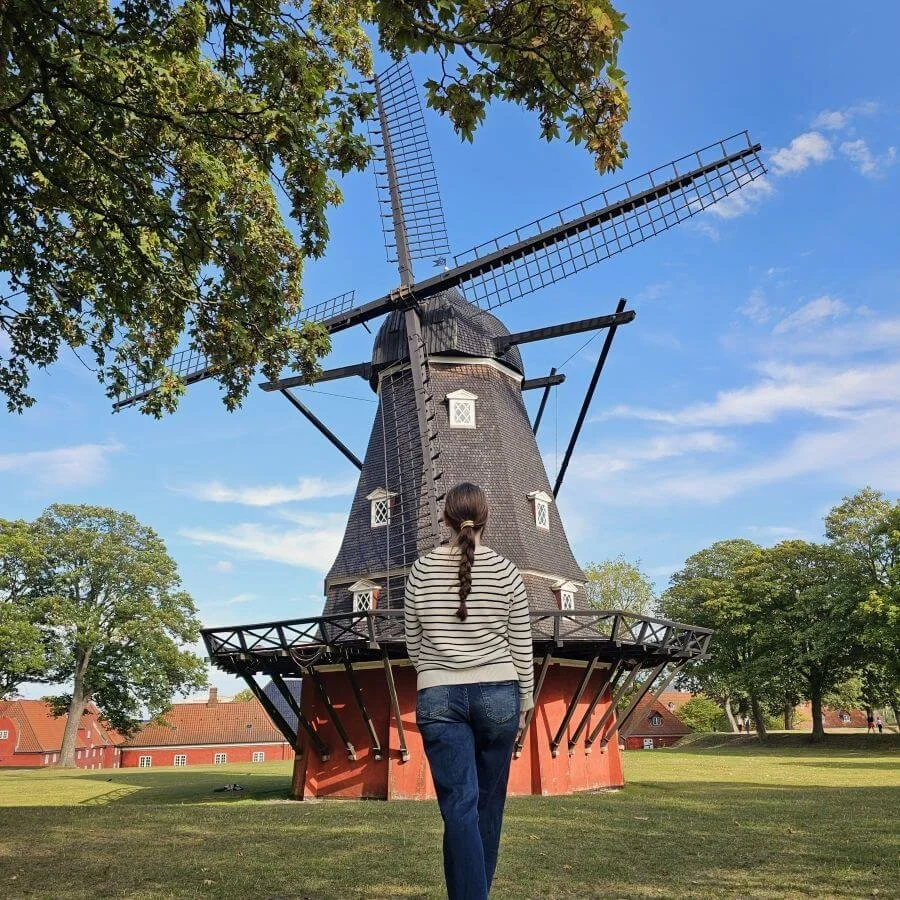 A woman in a striped sweater walks toward a windmill that is red on the bottom and black on top in Copenhagen.
