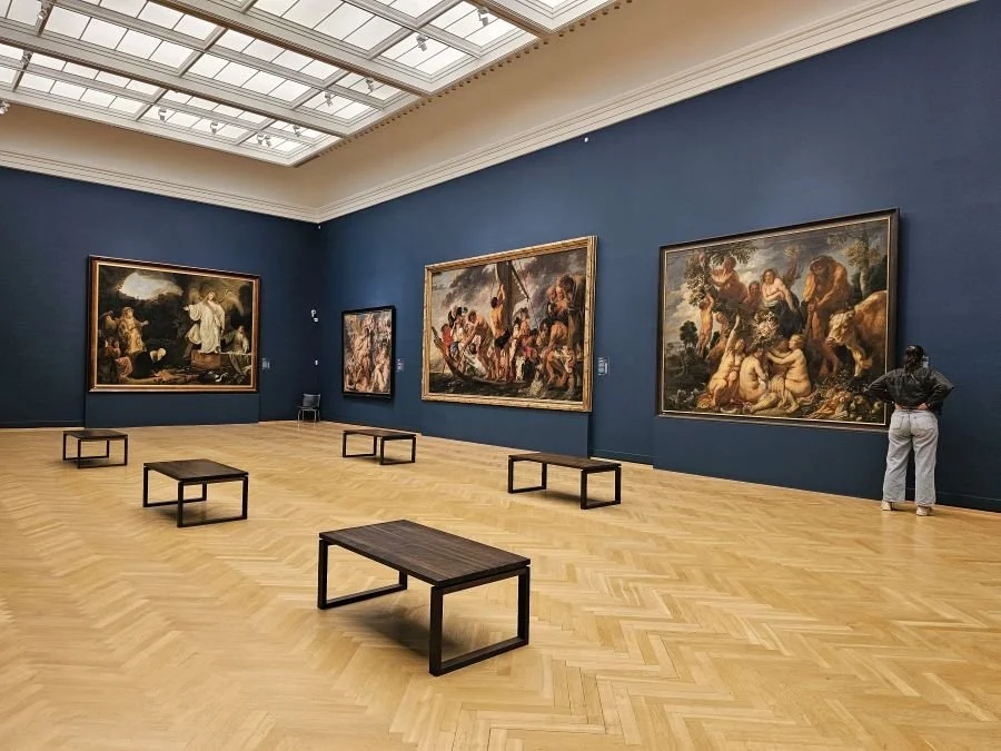 A dark blue room at a museum in Copenhagen with large painting hung on the wall and five benches in the middle of the room.
