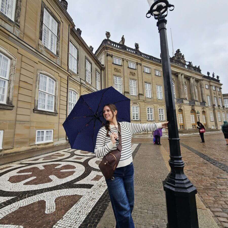 A woman in a striped sweater holds a blue umbrella in one hand with her other hand on a light post in front of Amalienborg Palace in Copenhagen.