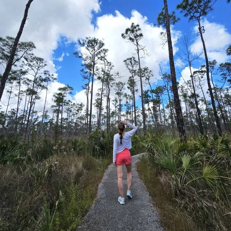 A woman in a white shirt and red shorts walks away from the camera on a paved trail through a pine forest