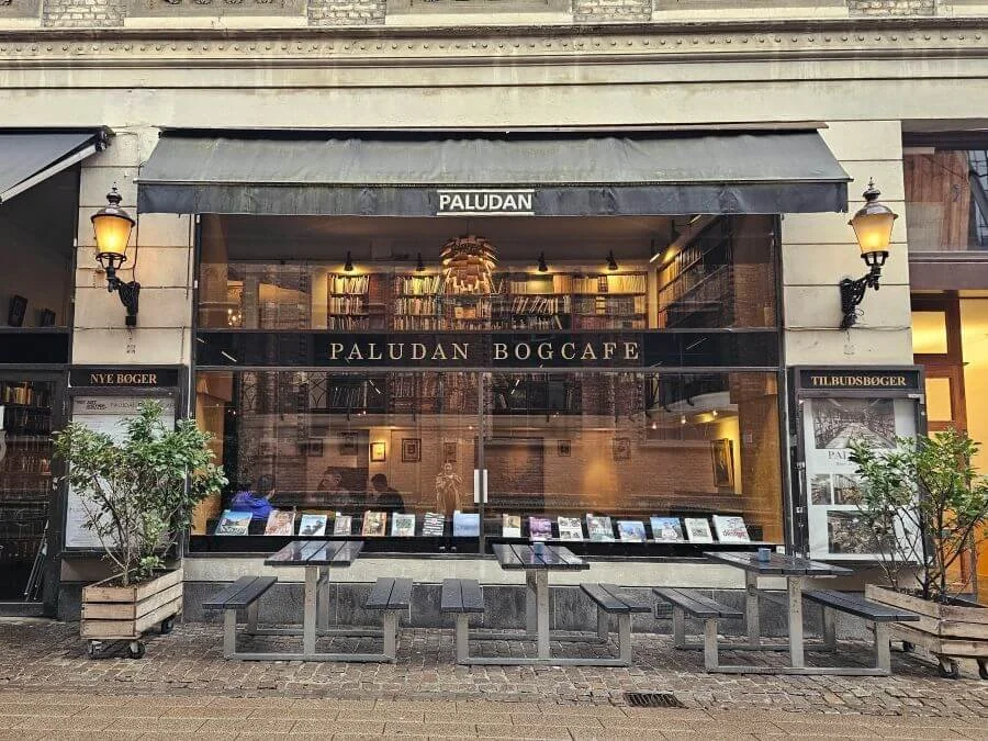 The storefront of a bookstore cafe, with floor to ceiling bookshelves behind a wall of glass windows as seen from outside looking in.