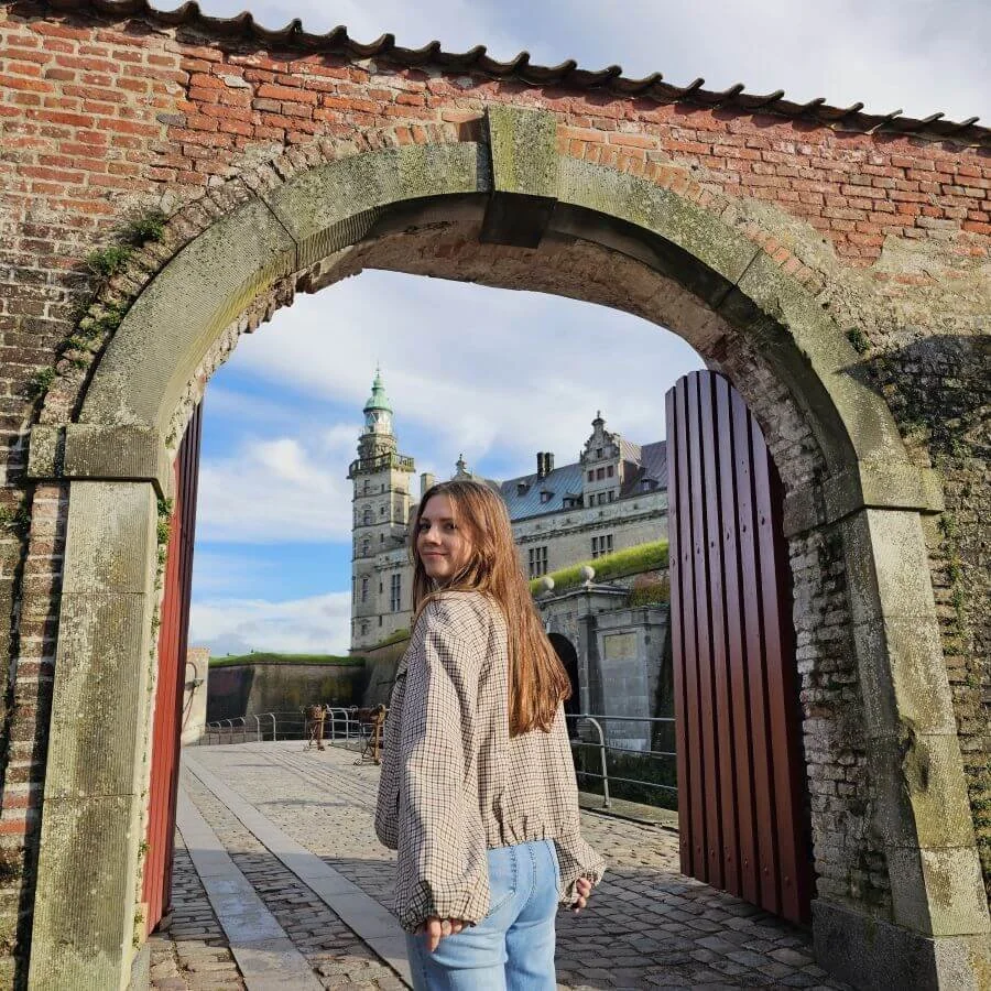 A woman in a plaid jacket stands under an arched stone doorway leading to the Kronborg Castle with a green turret in the background.
