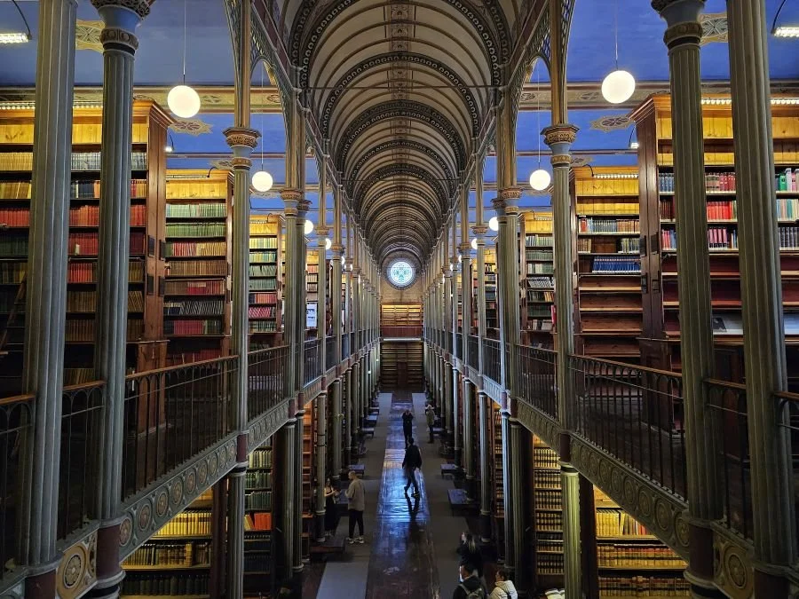 A huge historic library, with a view looking down the middle with several alcoves of bookshelves on each side on first and second floors.