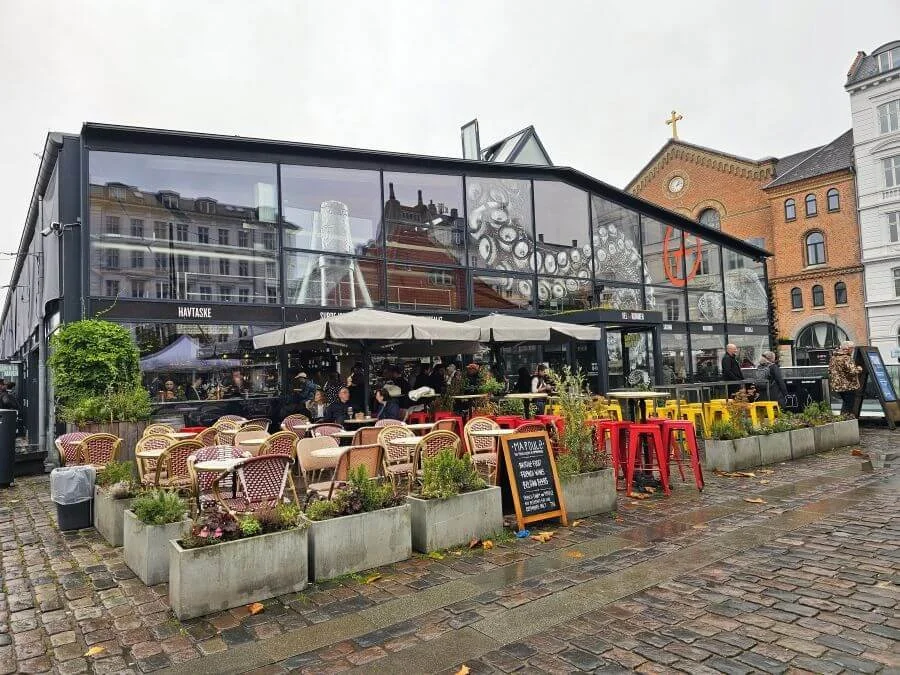 A glass walled food hall in Copenhagen with small tables and chairs under umbrellas in the front.