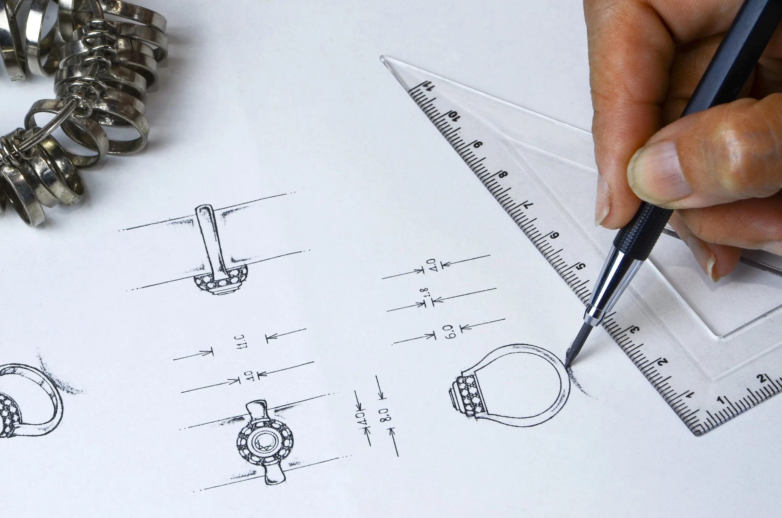 Hand drawing technical design schematics of jewelry, with a ruler and caliper on a white workspace.