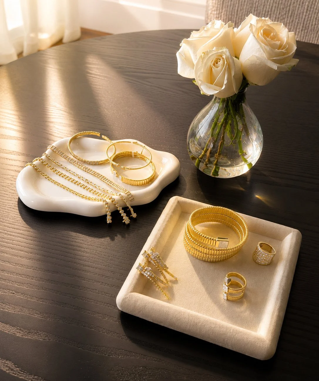 Gold and diamond jewelry sit in a display layout on a wood table with a vase of white roses.