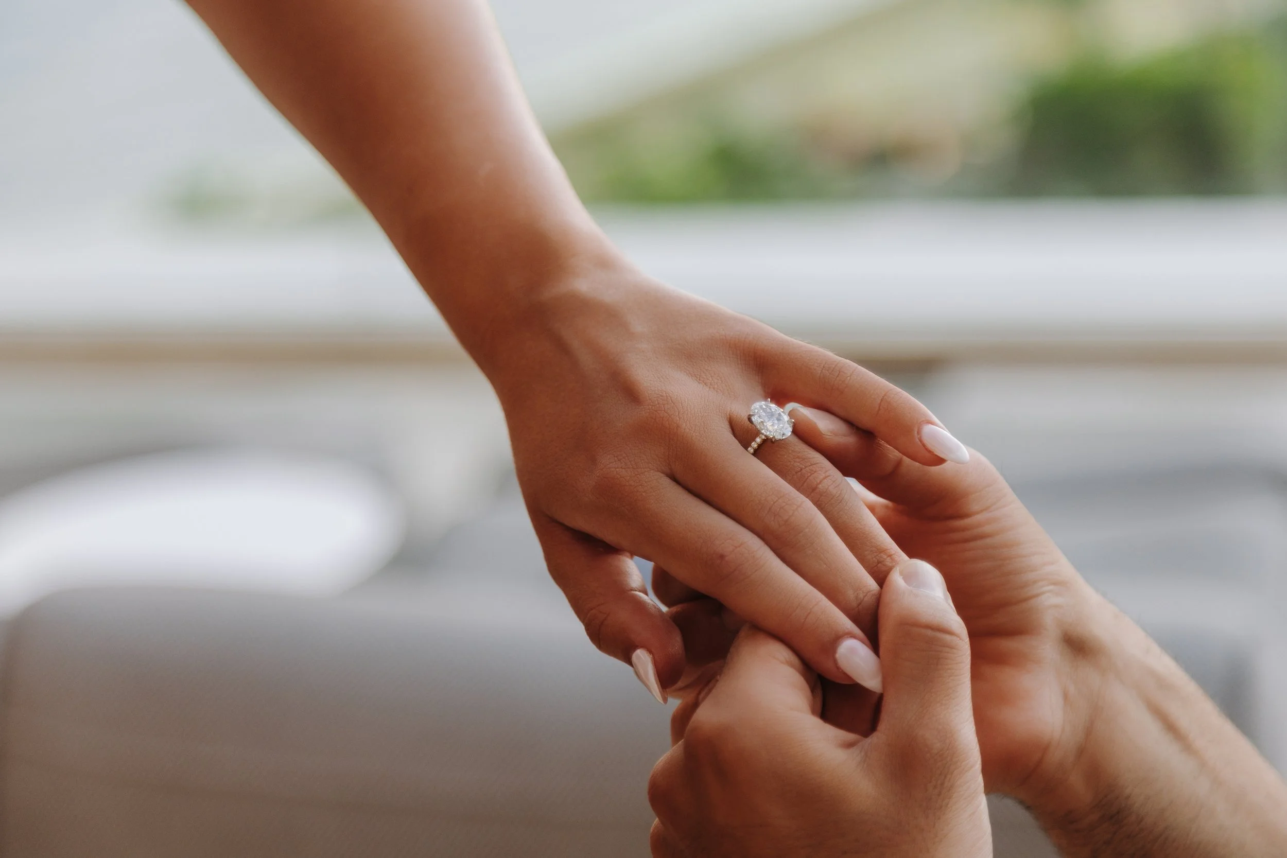 Close-up of a woman's hand with an engagement ring, holding a man's hand. They are touching, and the woman's hand is resting on the man's hand, with a blurred background.