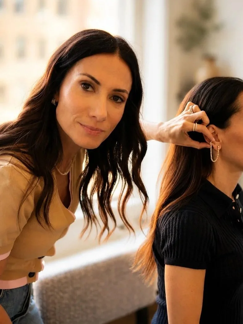 A woman with dark wavy hair is looking at the camera while adjusting earrings on another woman with straight, brown hair, who is turned to the side and wearing a black top.