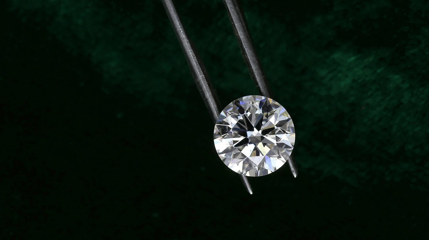 Close-up of a single round-cut diamond held by black tweezers against a dark green background.