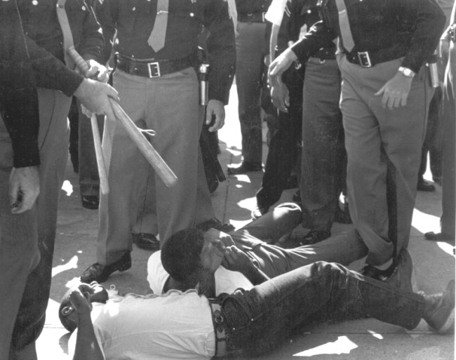 While FBI agents stood by, SNCC volunteers were beaten and arrested by sheriff’s deputies for attempting to bring water to people (many elderly) waiting in the hot sun for hours to register to vote in Selma, 1963. Image: © John Kouns.