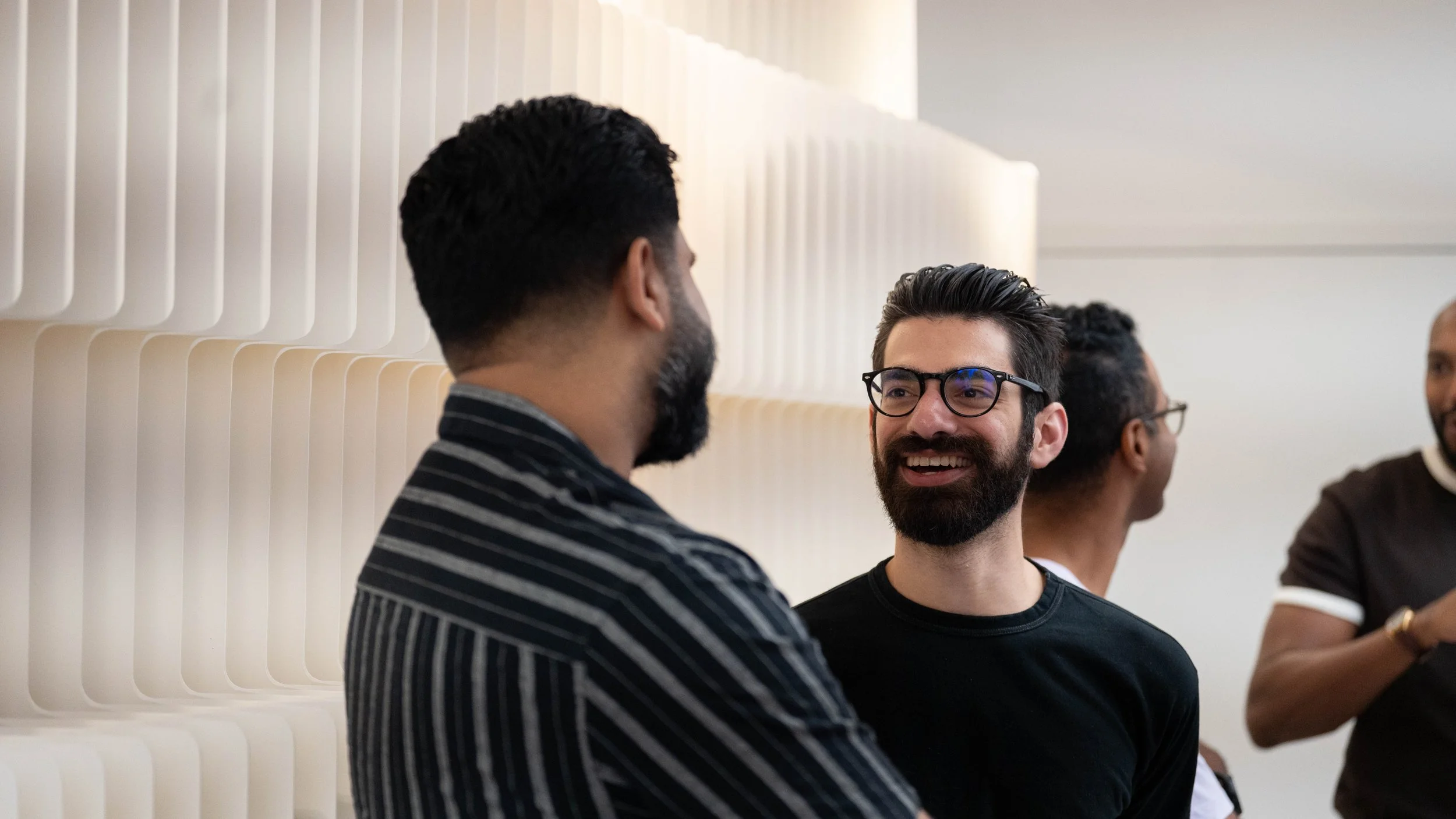 A group of diverse people talking and smiling, one person is wearing glasses, in a modern indoor setting with a light-colored wall.