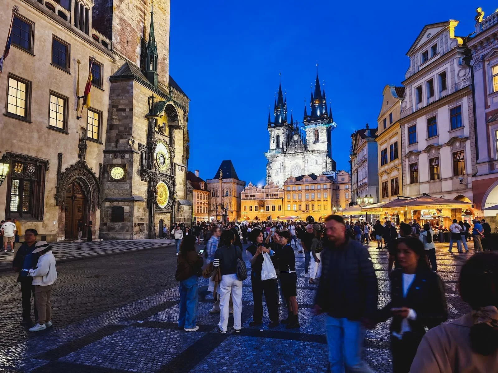 Illuminated historic buildings and Gothic towers of the Church of Our Lady before Týn in Prague's Old Town Square at blue hour.