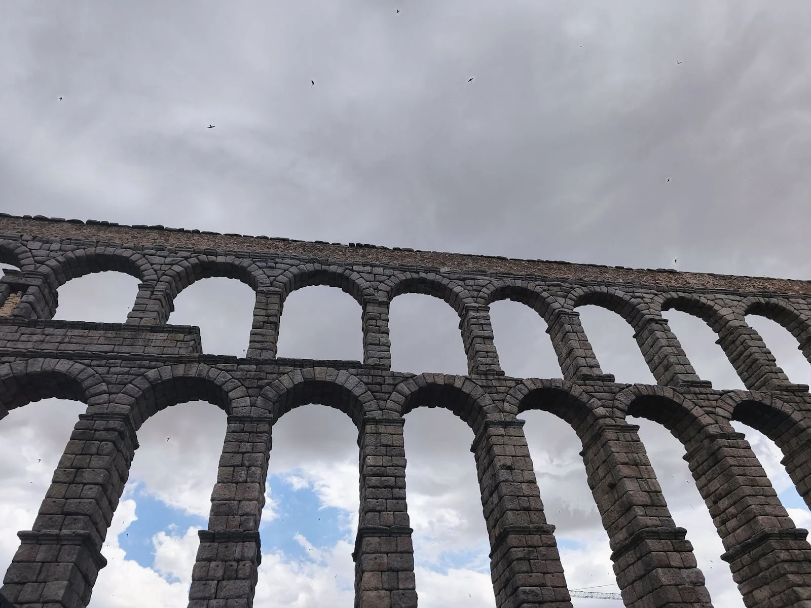 Roman Aqueduct of Segovia seen from below, with stone arches against a cloudy sky.