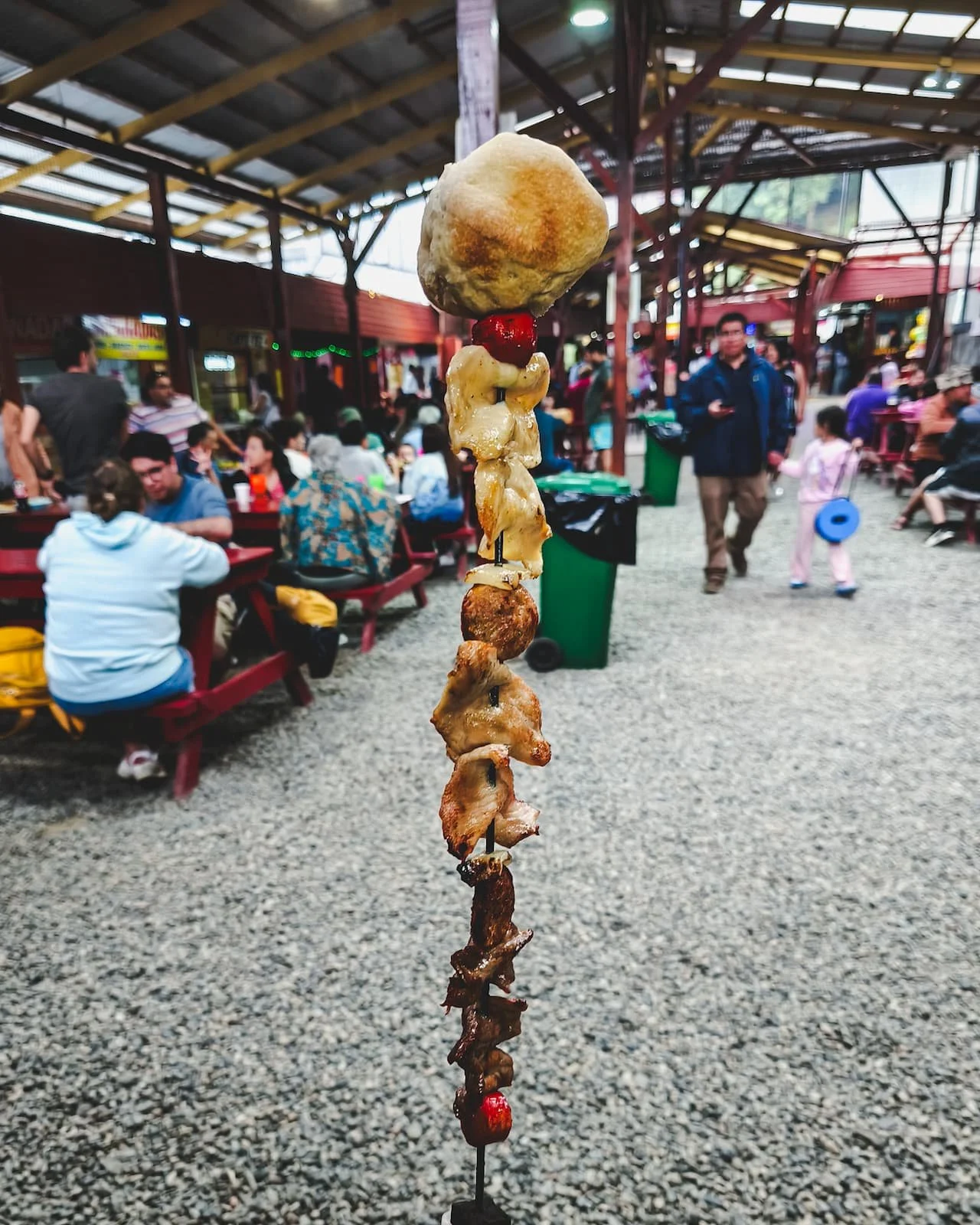 Large anticucho served at Feria Encuentro Costumbrista with food stalls and seating in the background.