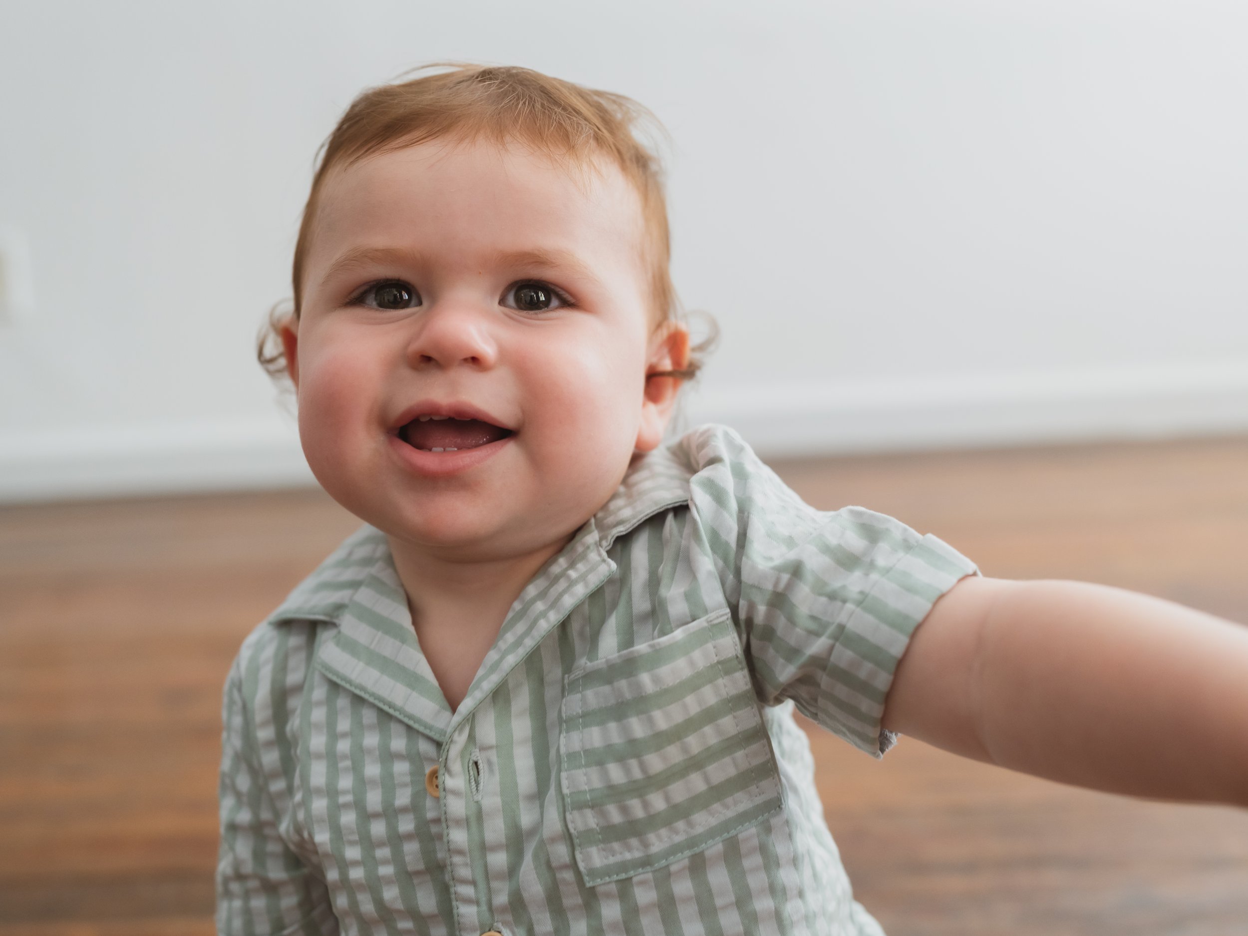 A smiling young child with red hair, wearing a light gray and white striped shirt, reaching out toward the camera, on a wooden floor with a plain white wall in the background.