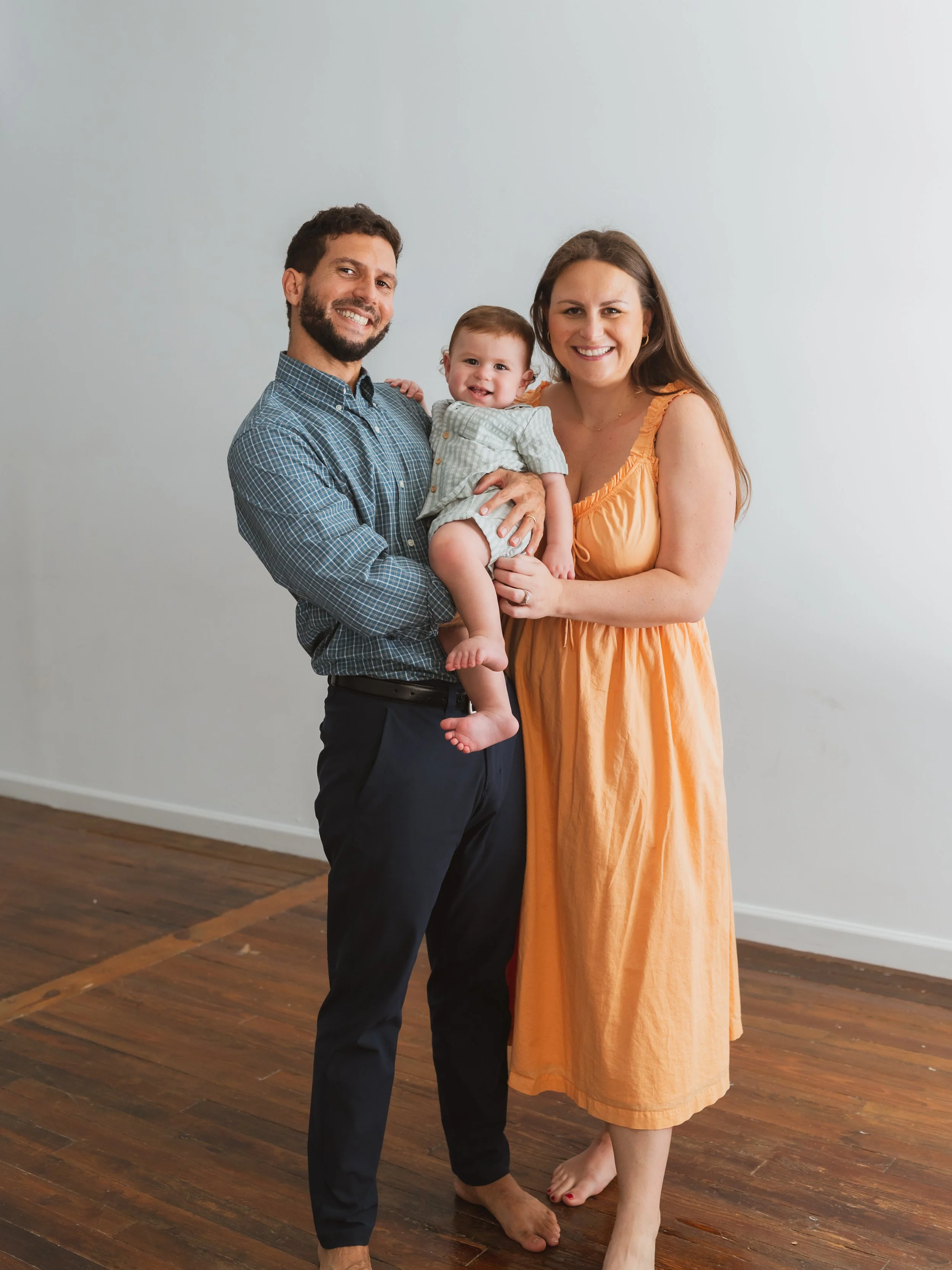 A happy family of three standing indoors on a hardwood floor, smiling at the camera. The father is holding a young child, and the mother stands next to them in an orange dress.