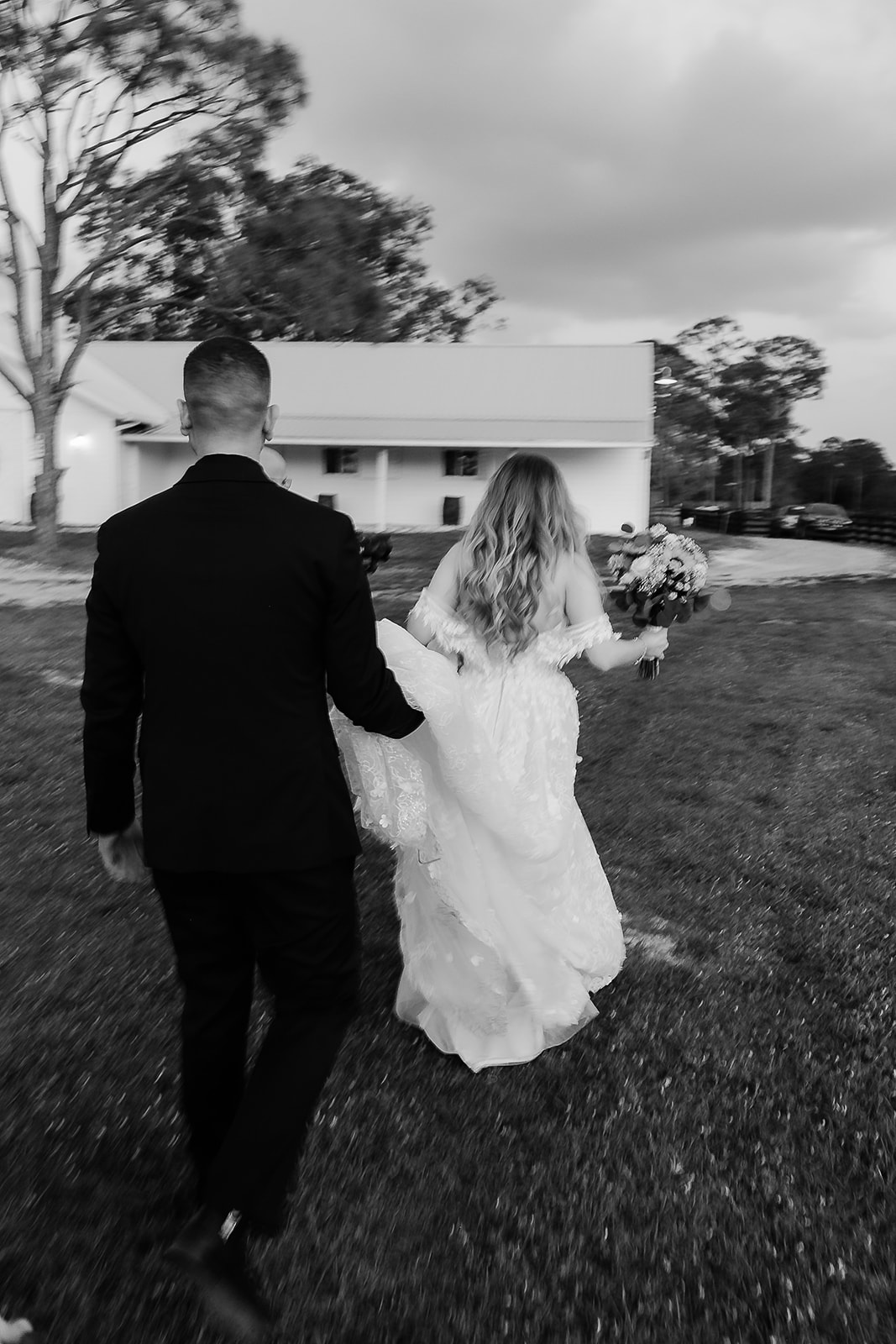 A bride in a wedding dress holding a bouquet of flowers walking with a groom in a black suit on a grassy area outside between trees and a building.