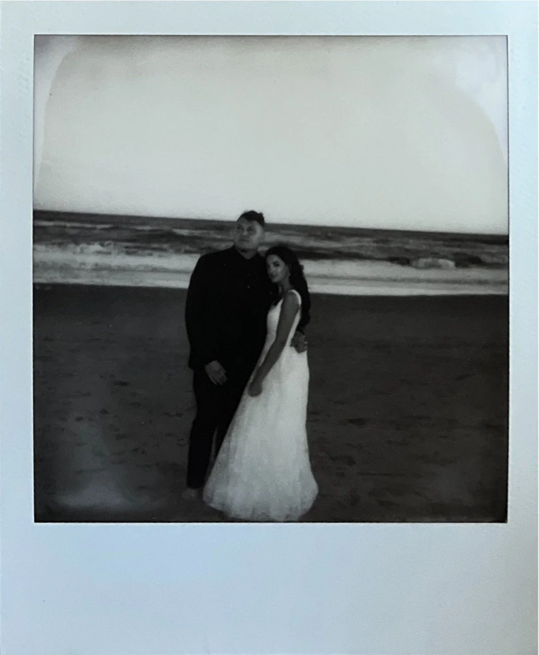Black and white photo of a couple on a beach, with the man wearing a suit and the woman in a white gown, standing closely together.
