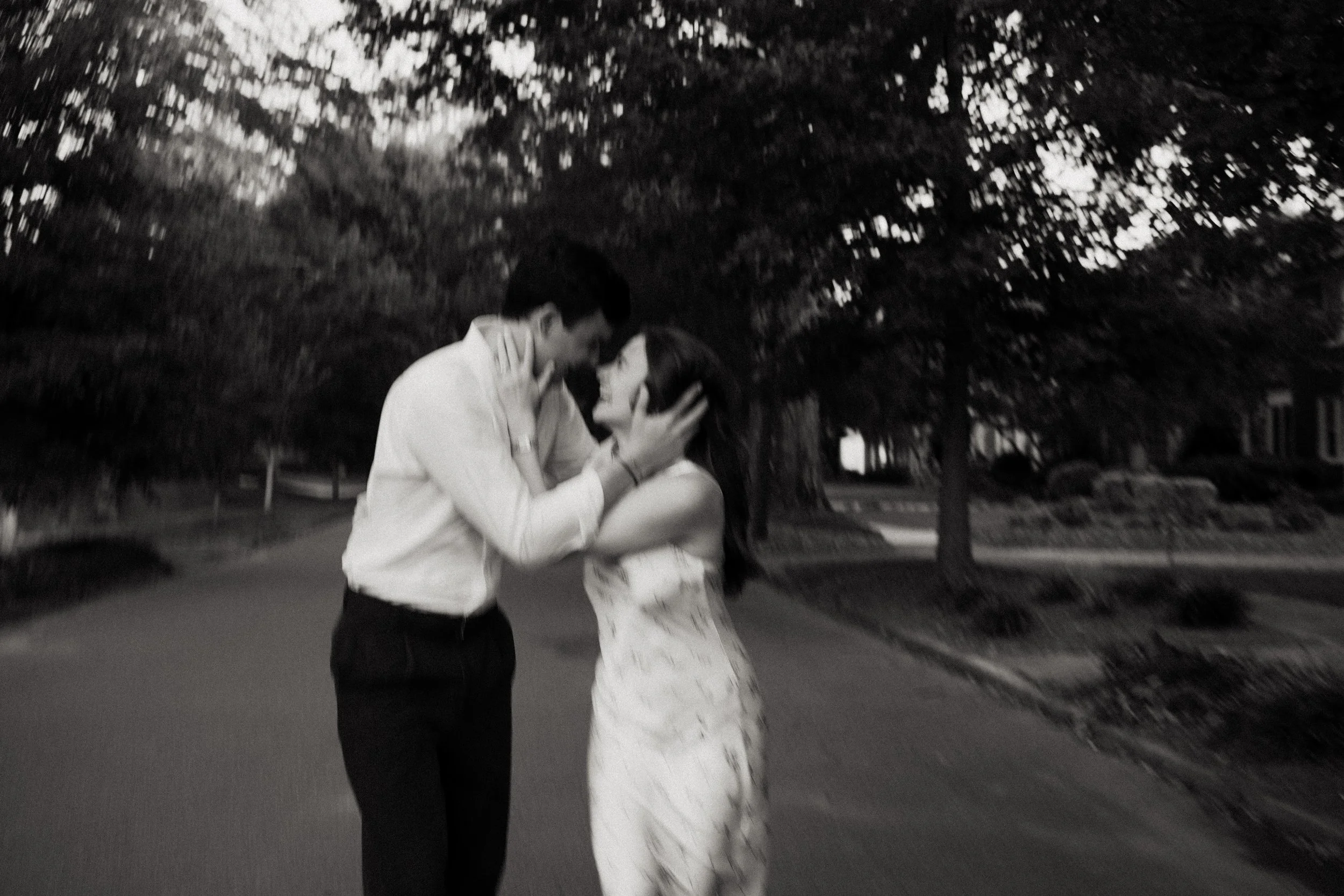 A black-and-white photo of a couple standing close together outdoors, with trees and a sidewalk in the background, sharing an intimate moment with their foreheads touching and hands on each other's faces.