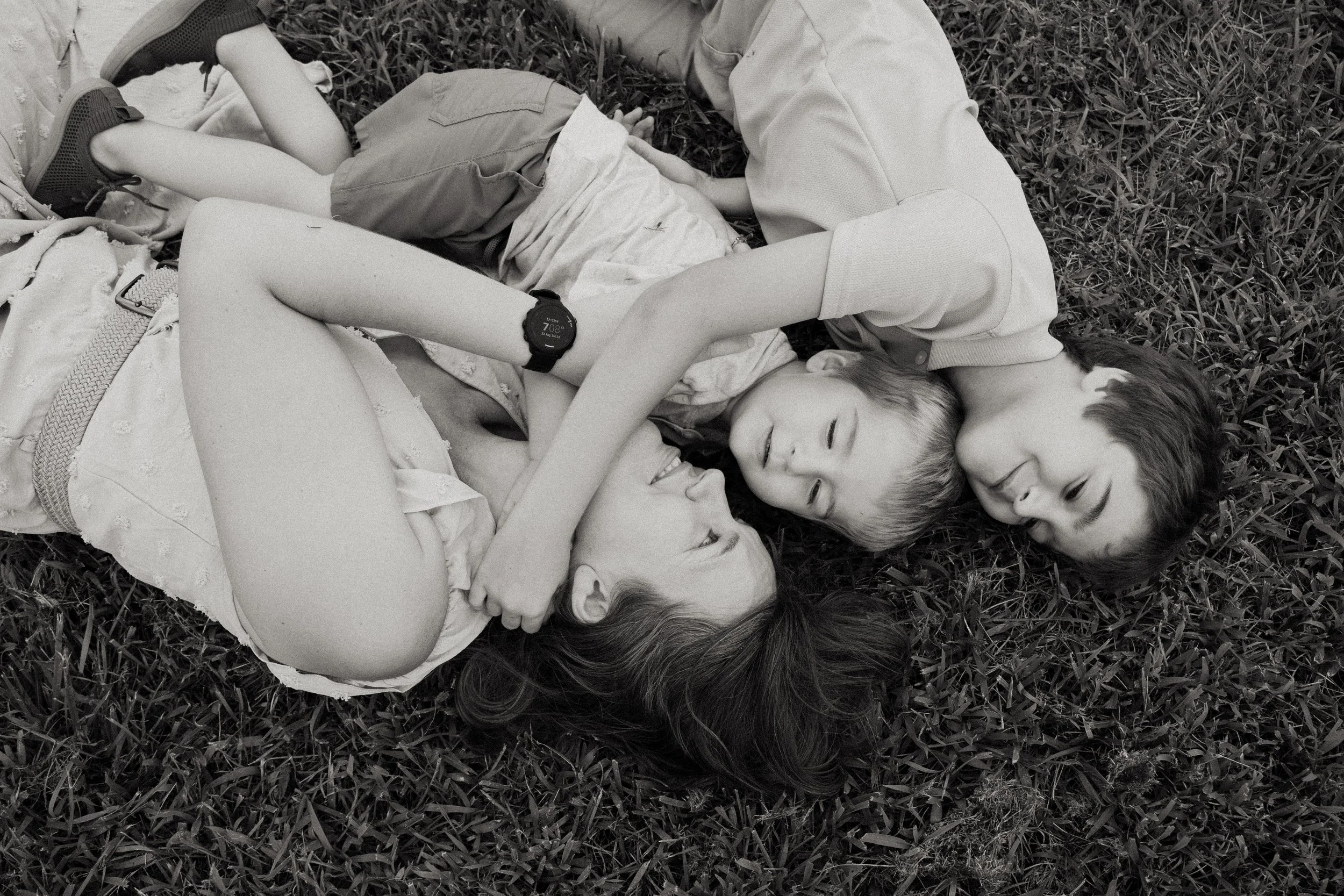 Three children lying on grass, two boys and one girl, engaging in playful interaction with the girl and one boy smiling and the other boy looking at her.