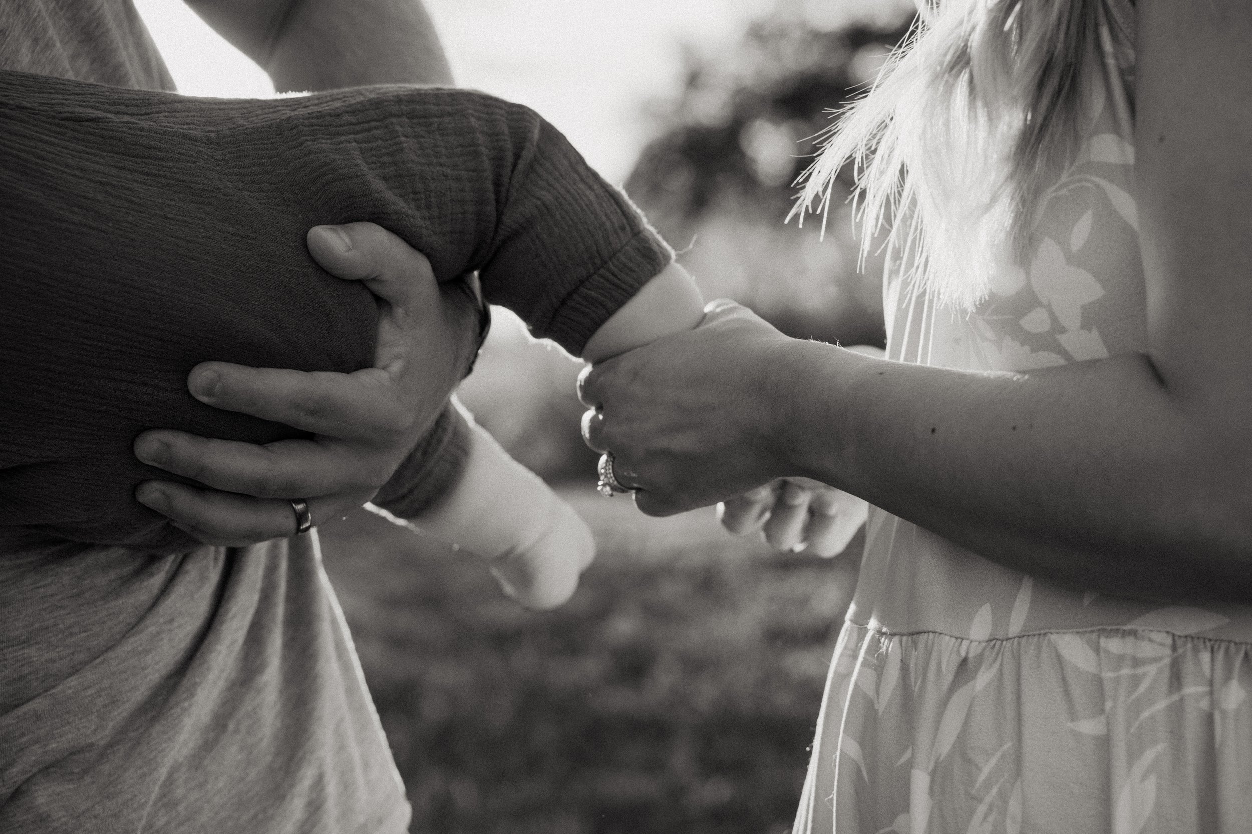 A couple exchanging rings outdoors, with a woman placing a ring on a man's finger. The black and white photo captures a close-up of their hands, emphasizing the hands and rings, with a blurred background.