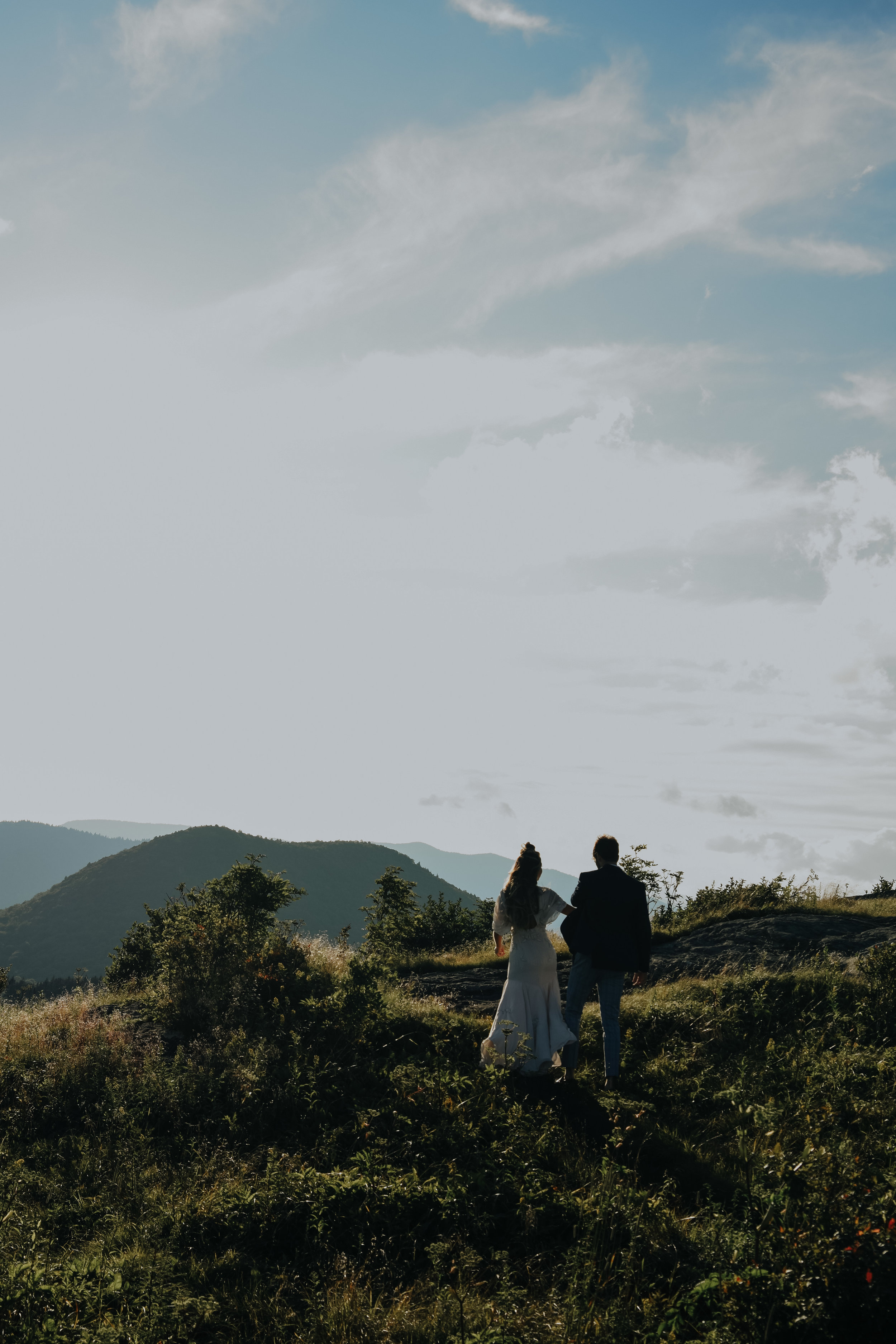  An image at a distance from behind, of a couple walking together after their elopement, along the crest of a grassy hill as they look out upon rolling hills and open sky at Black Balsam Knob 