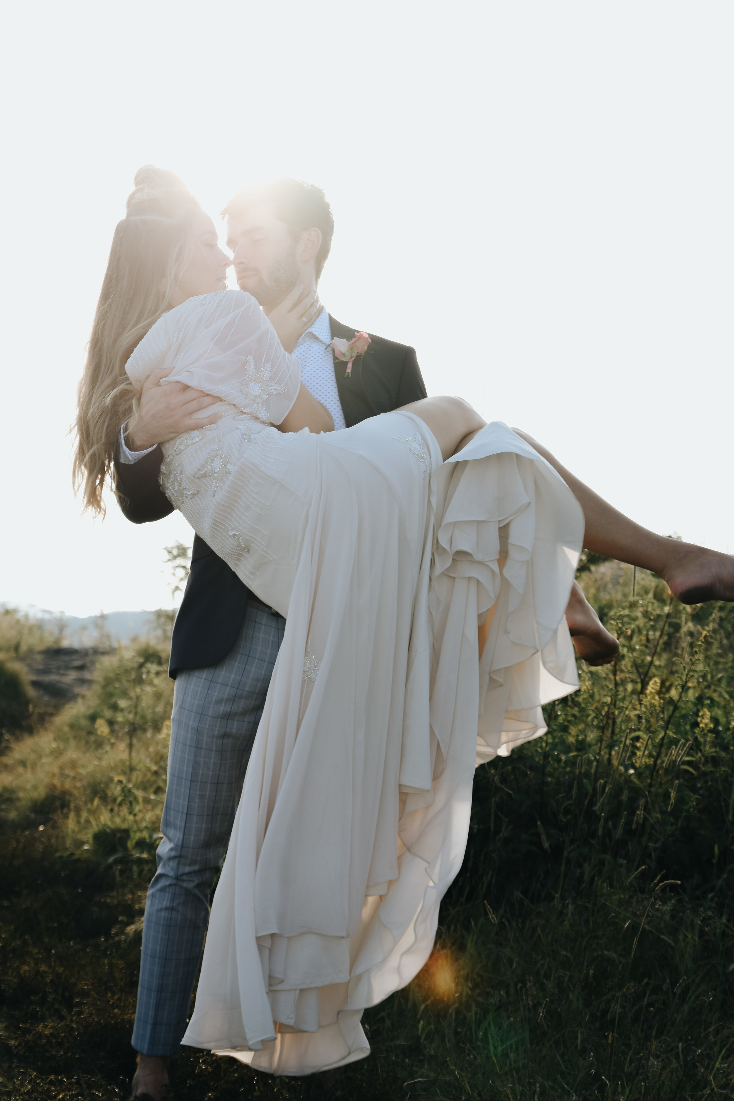  A picture of a couple who have eloped, the groom romantically lifting his partner up in his arms as they almost kiss, while the sun shines brightly between them by Vanessa Venable - Asheville North Carolina elopement photographer 