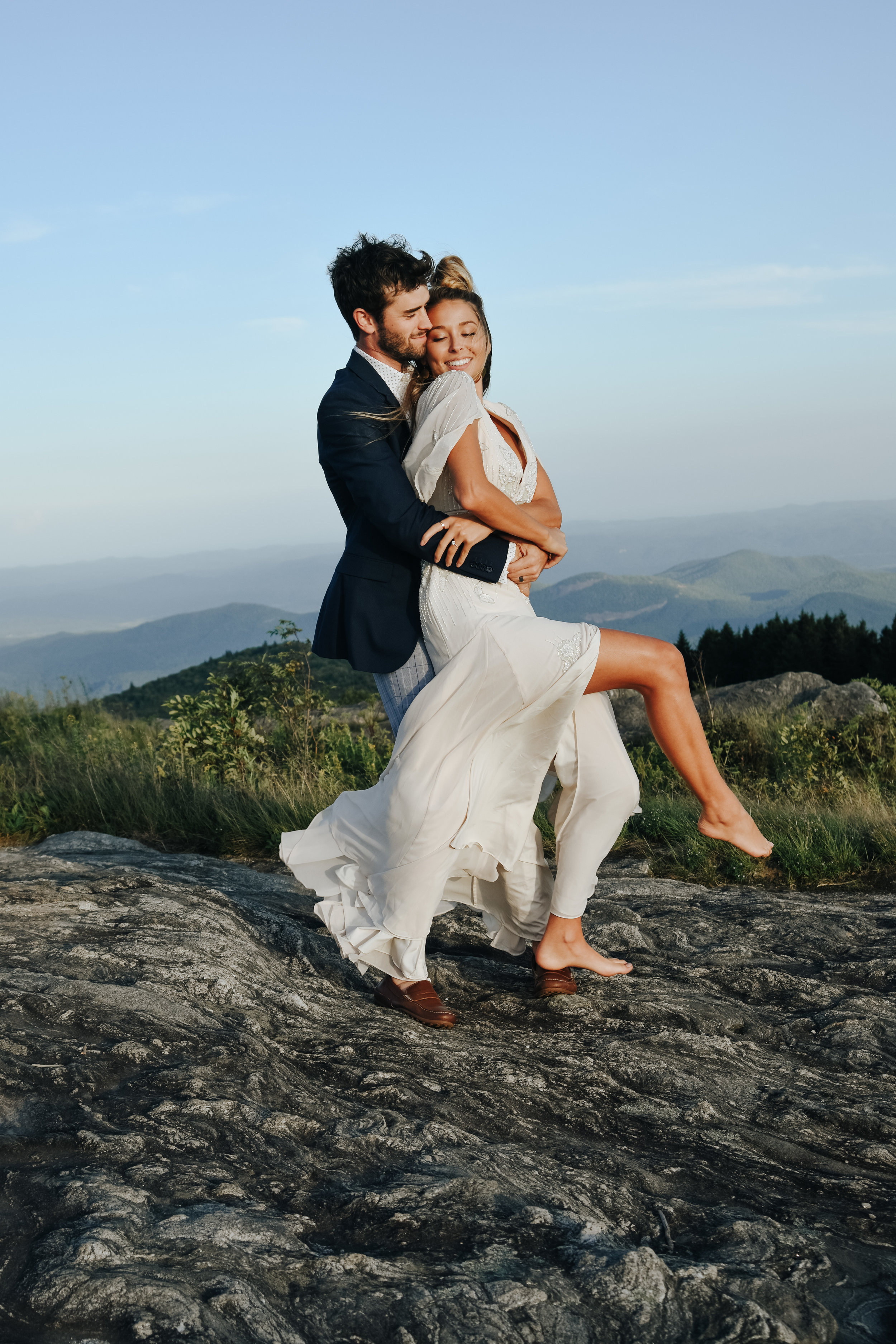  A photograph of a couple who have just eloped, with the groom holding his partner from behind as she leans back against his chest, holding him tight as they stand high on a hill that overlooks mountains beyond at a Black Balsam Knob elopement 