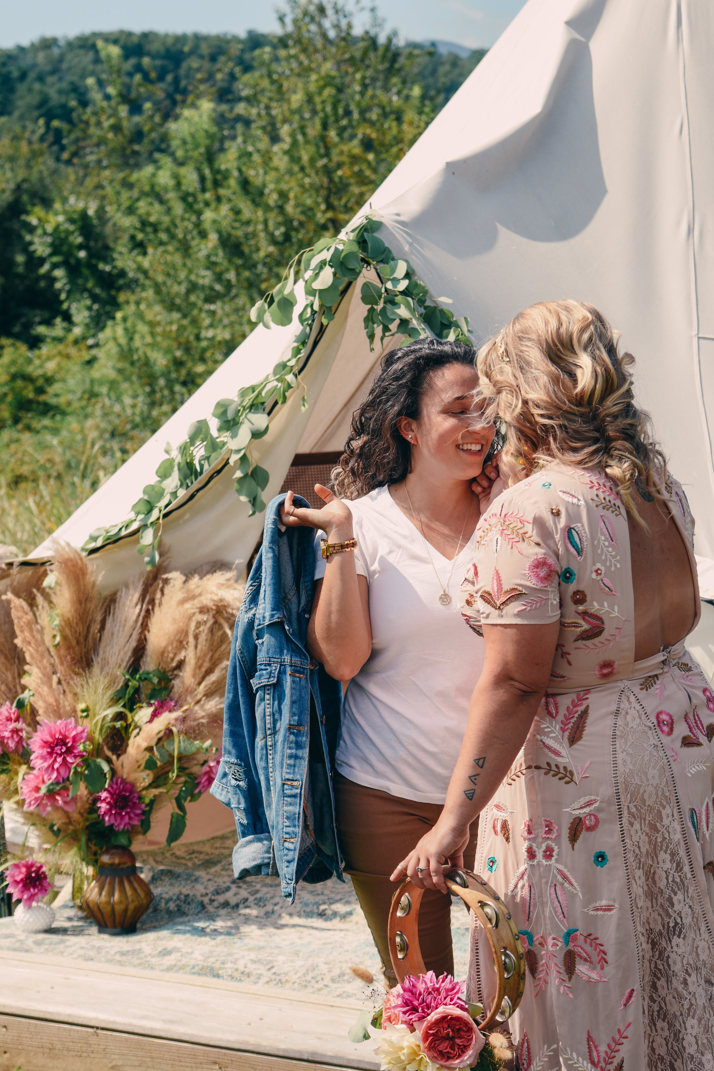  An LGBTQ+ couple stand in front of their rustic teepee with drive flower arrangements and the mountain-side greenery in the background  