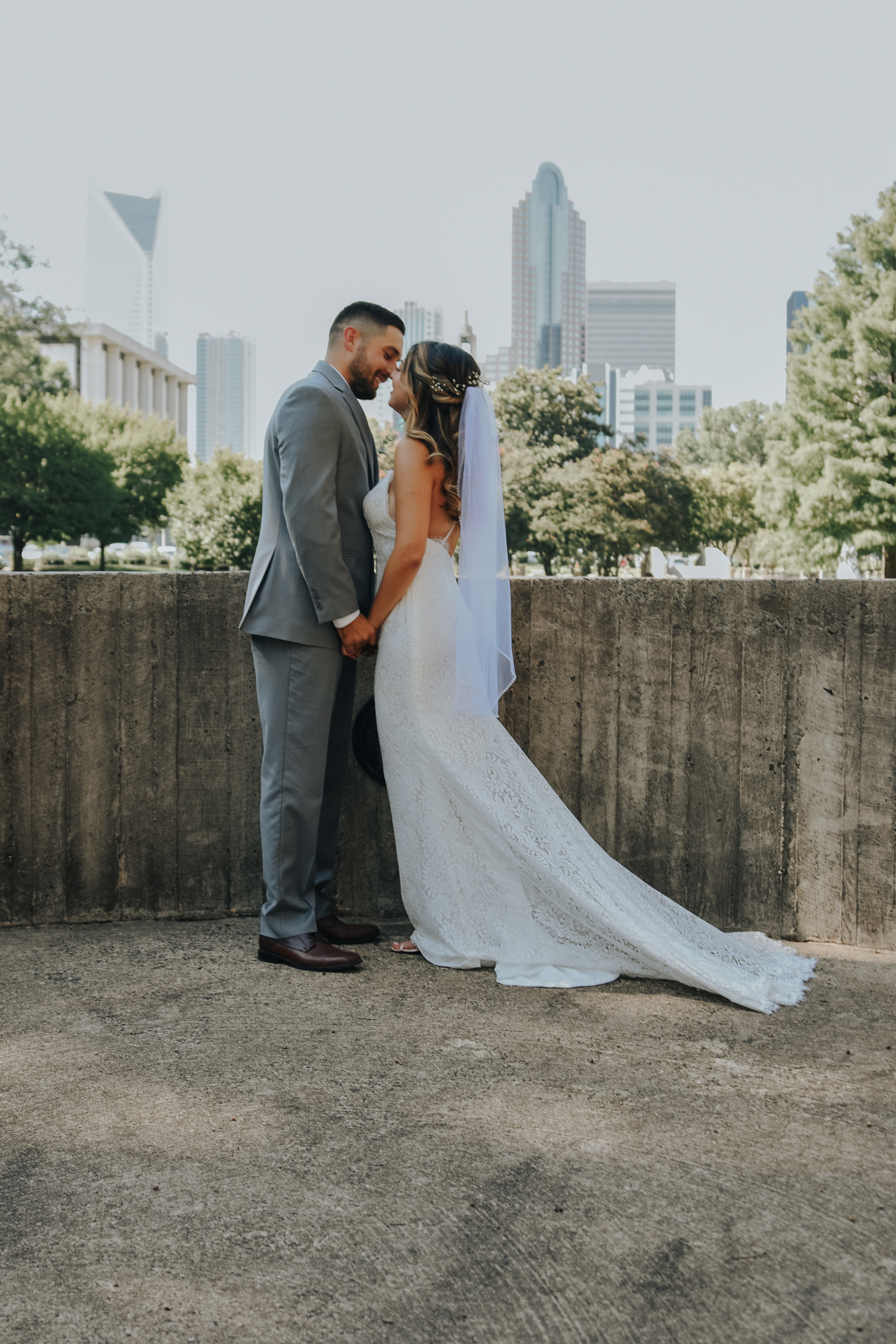  A picture of a couple celebrating their elopement, standing lovingly close together, almost kissing, as they hold hands with the city skyline behind them in Marshall Park by Vanessa Venable - Charlotte North Carolina elopement photographer 