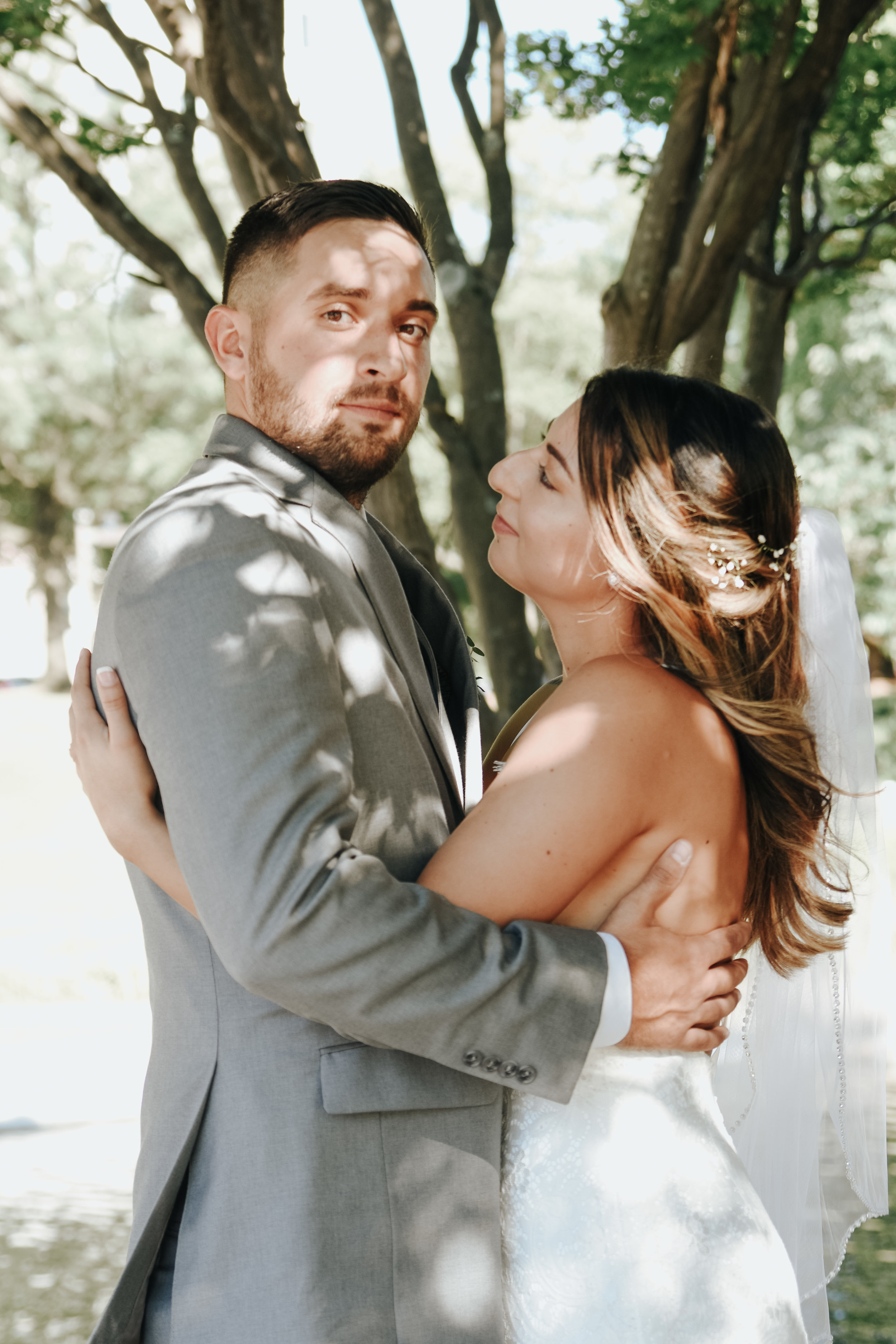  A photograph of a bride and groom tenderly embracing shortly after their elopement as they stand outdoors under a tree in the sunlight and shadows Mecklenburg County Courthouse elopement 