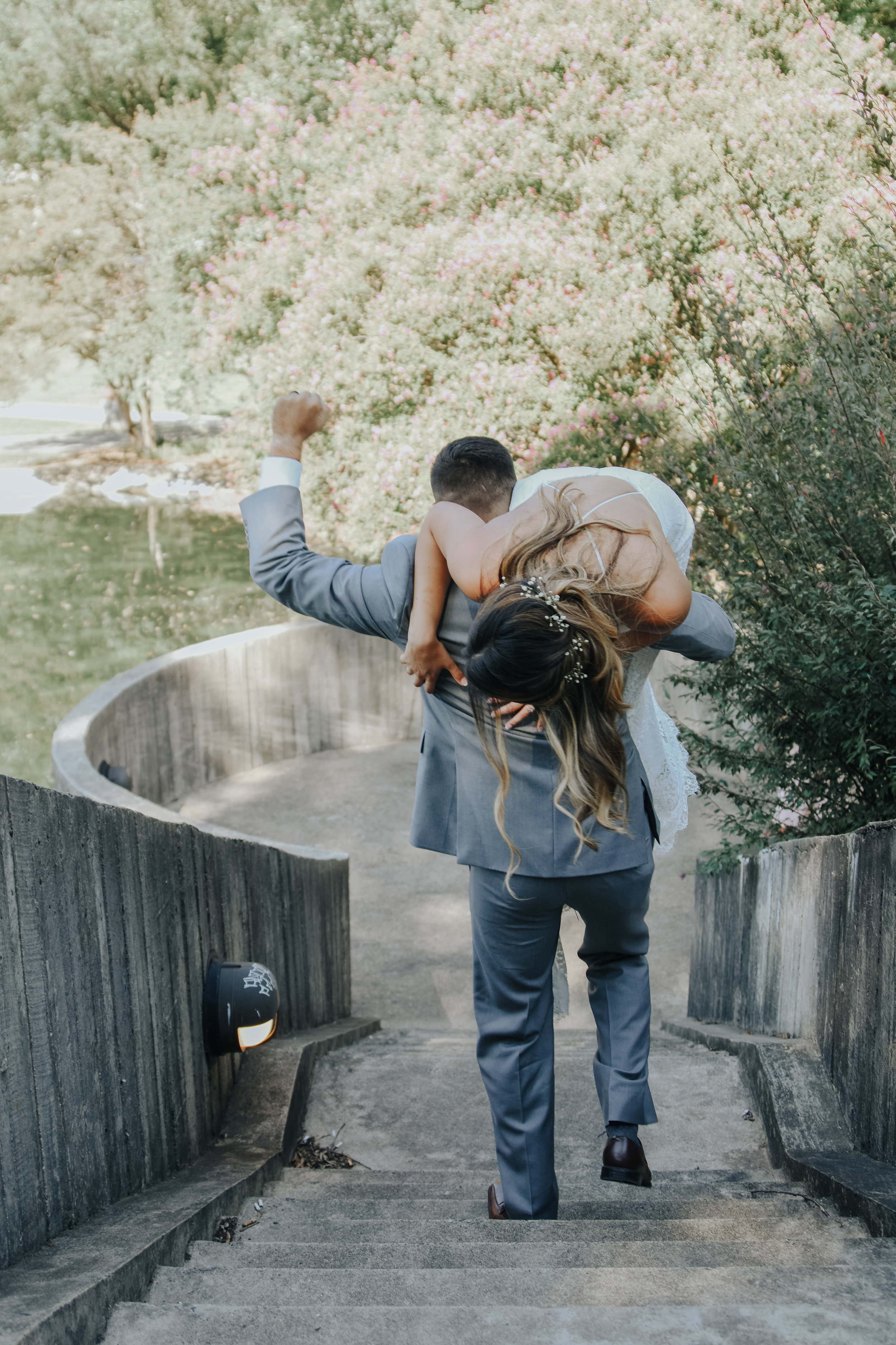  A photograph of a couple who just eloped, walking down some stairs outdoors as the groom playfully holds his partner over his shoulder, raising a happy fist in celebration Mecklenburg County Courthouse elopement 