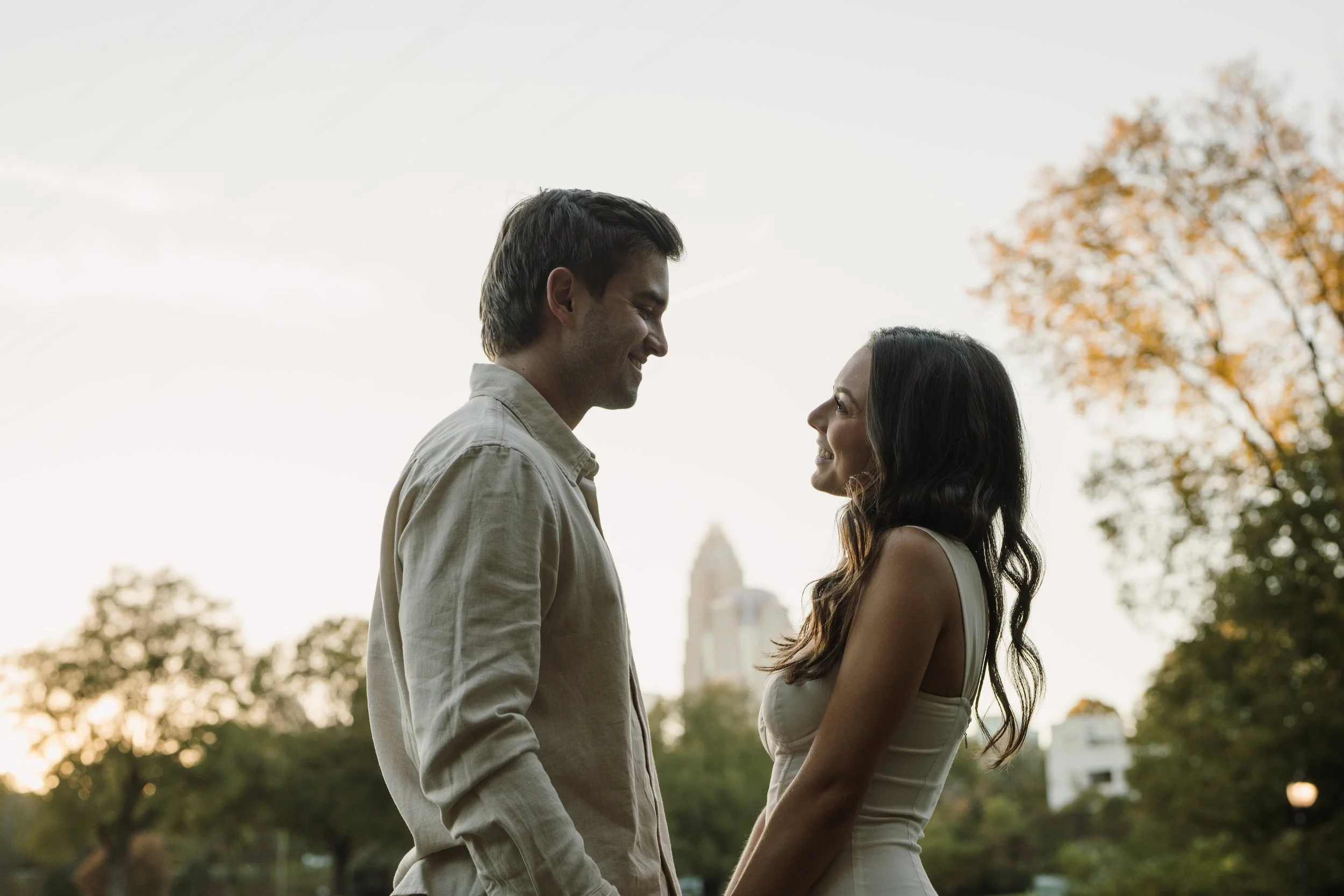 A man and woman stand face-to-face outdoors, smiling at each other during sunset, with trees and city buildings in the background.