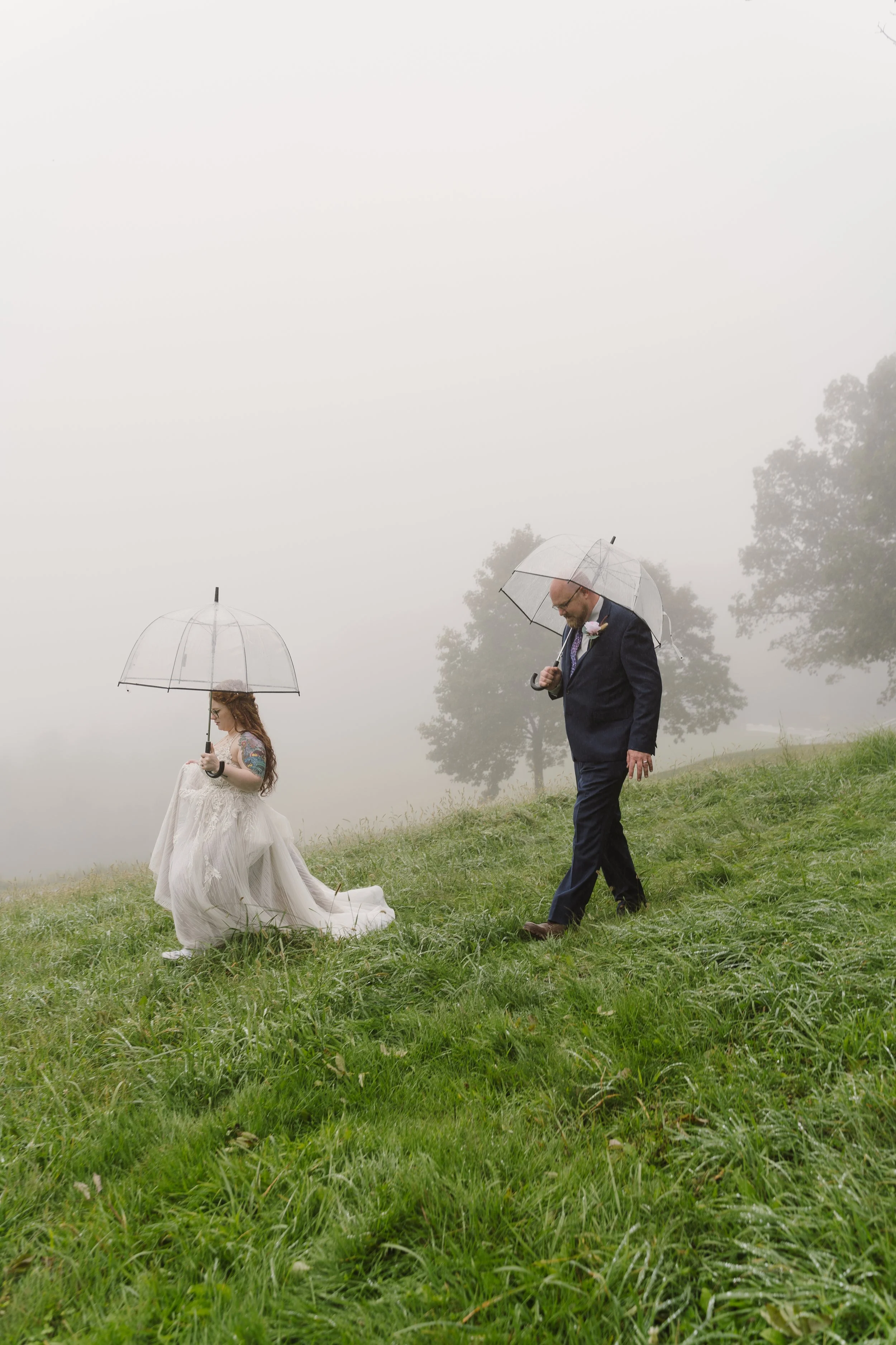 A bride and groom walking on a foggy grassy hill, each holding transparent umbrellas, with the bride dressed in a wedding gown and the groom in a dark suit.