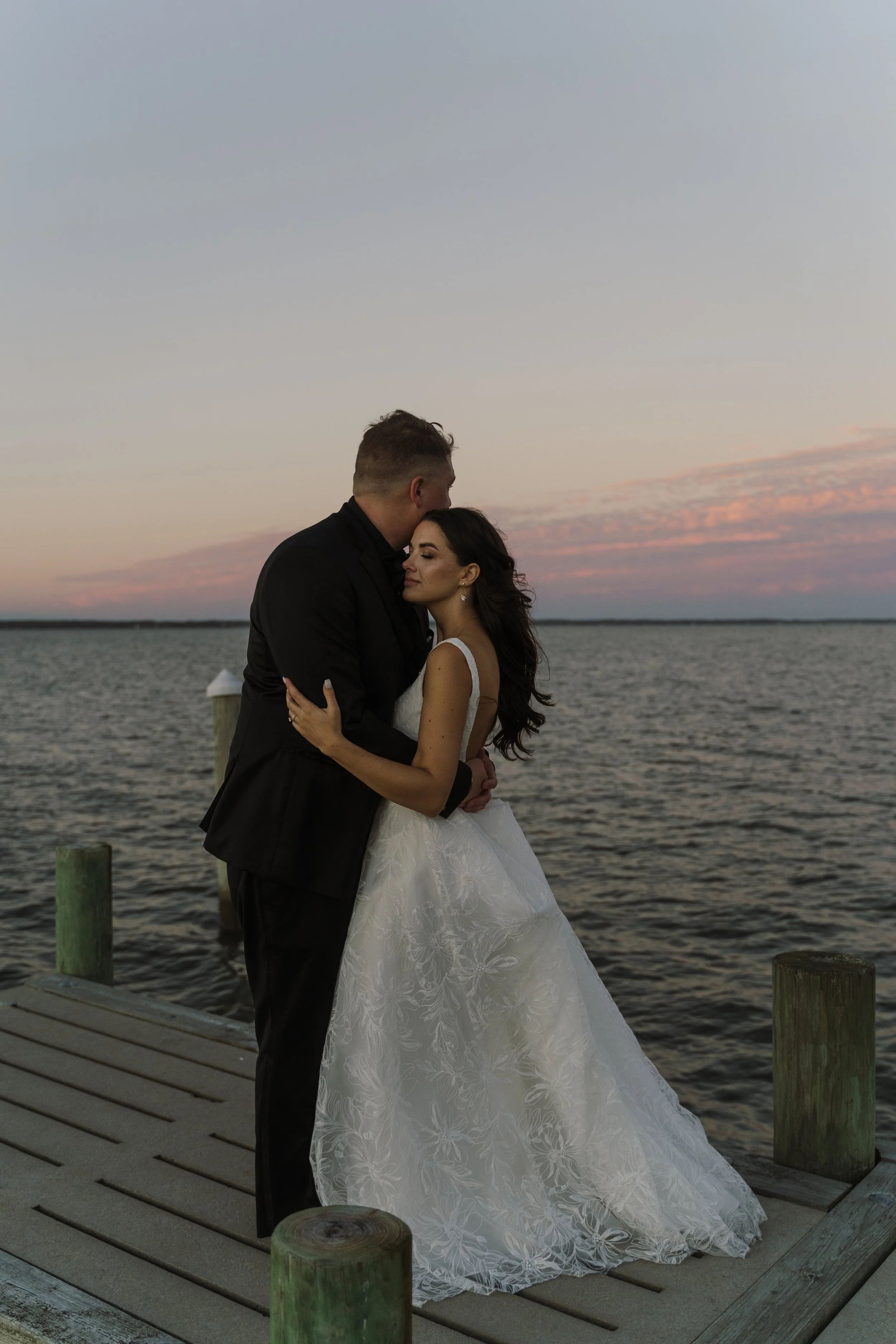 A couple in wedding attire embrace on a dock by a body of water at sunset, with pastel sky and pink clouds.