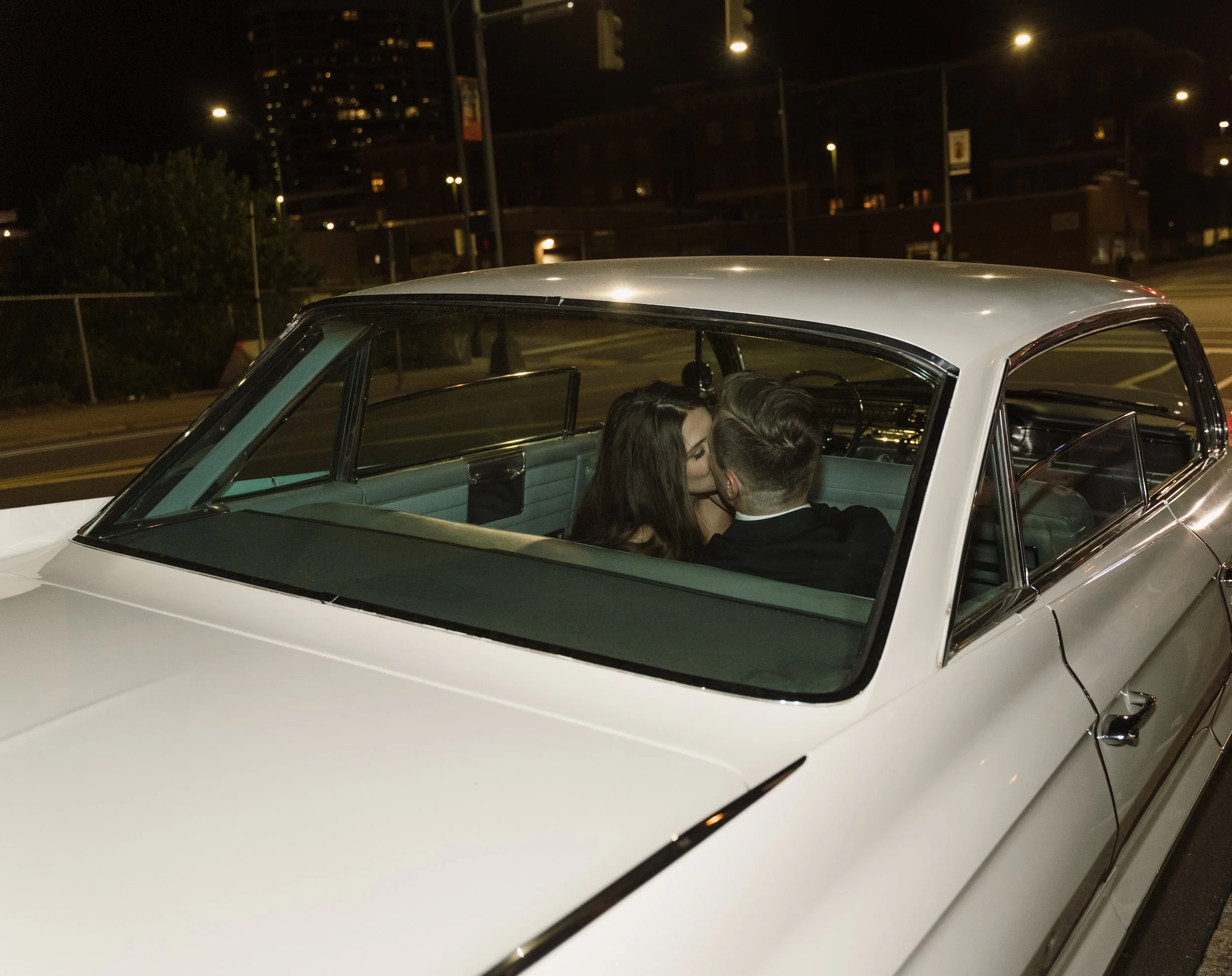 A couple sharing a kiss inside a vintage white car parked at night on a city street.