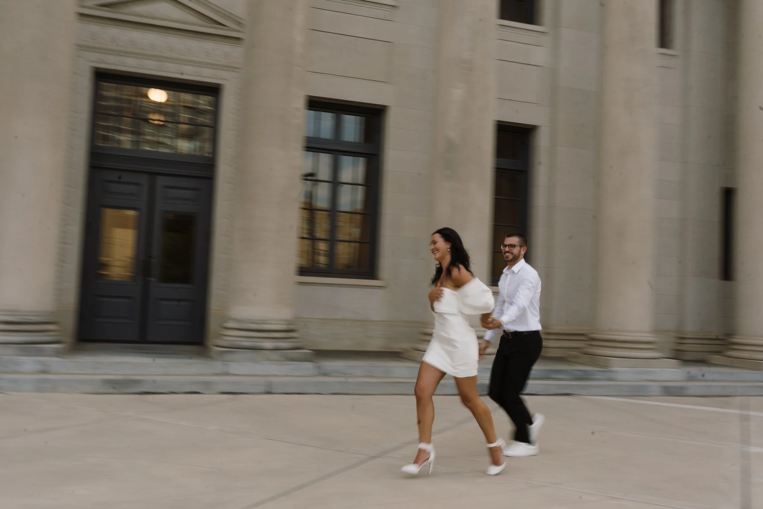 A couple smiling and holding hands as they run outside a large, historic-style building.