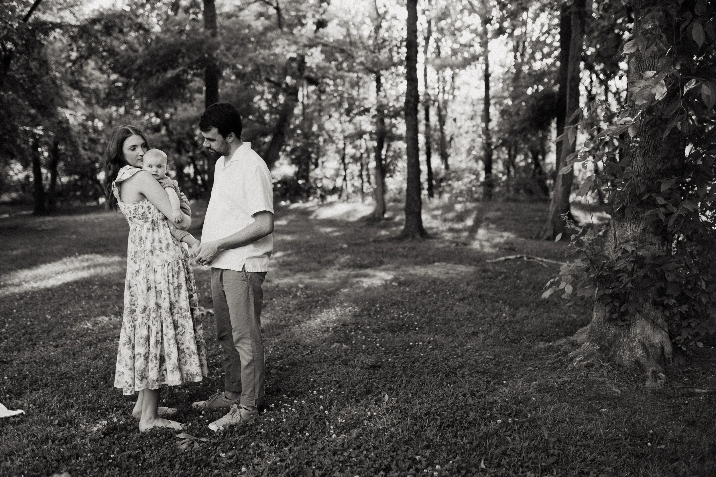 A family of three outdoors in a park, a woman holding a baby and a man holding her hand, with trees surrounding them.