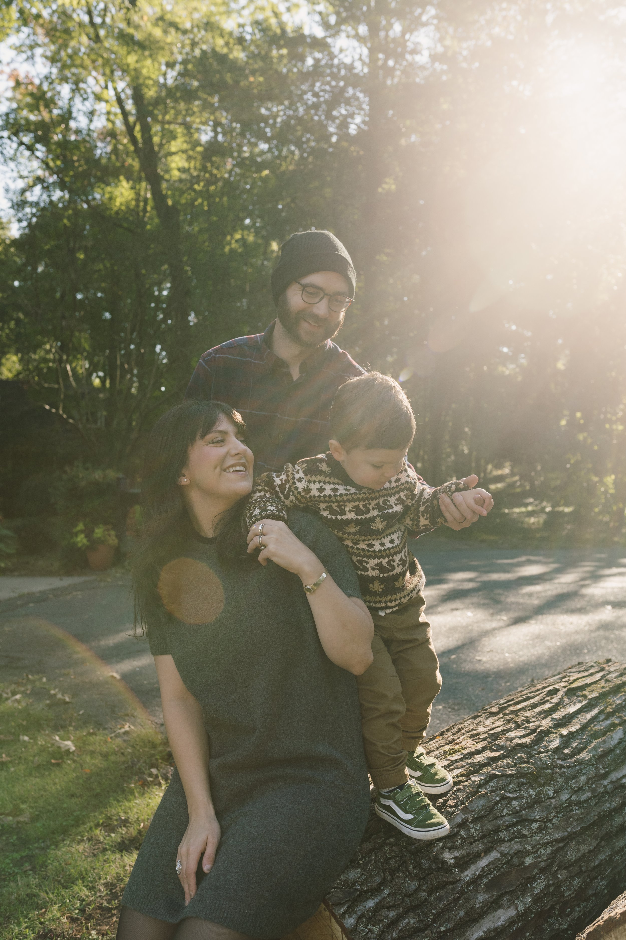 A family of three enjoying time outdoors in a park on a sunny day, with a woman sitting on a fallen tree, a man and a young boy standing beside her, the boy reaching out towards the tree and the woman smiling at the boy.