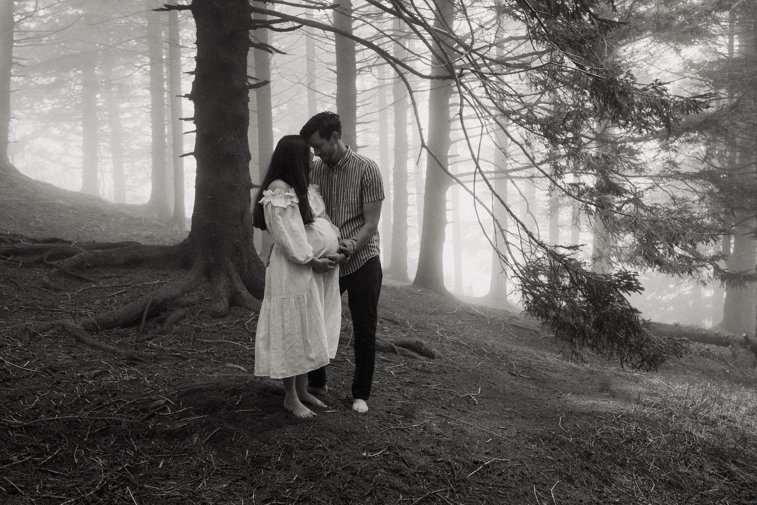 A black and white photo of a couple standing in a foggy forest, with the woman showing a pregnant belly and the man gently holding her belly, both with their heads bowed in a tender moment.