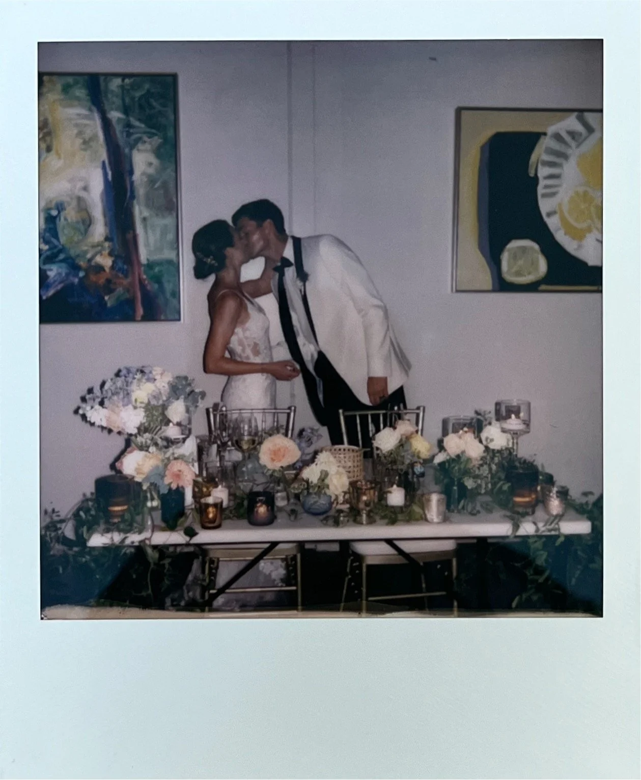 A bride and groom kissing at a wedding reception table decorated with flowers and candles, with abstract paintings on the wall behind them.