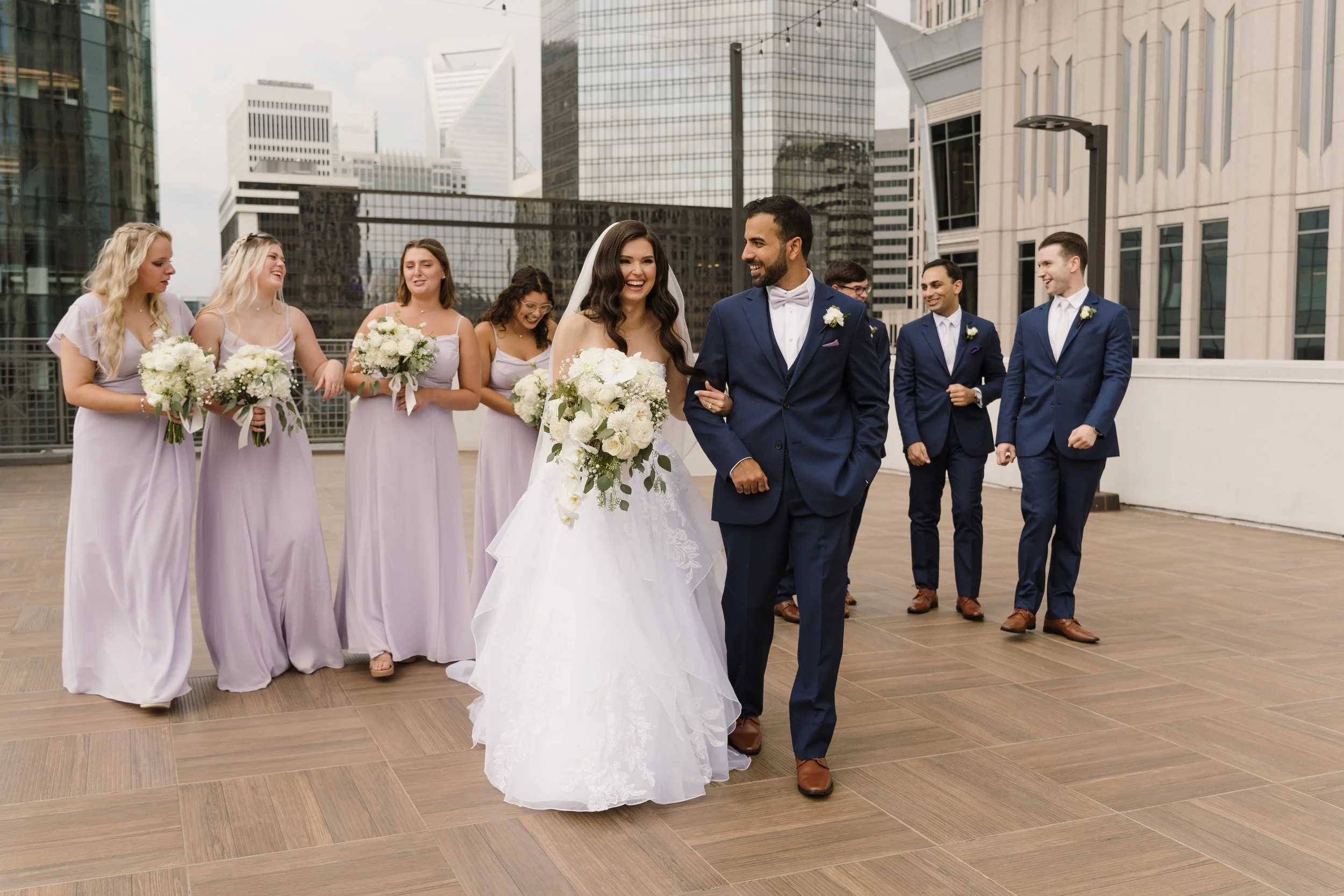 A bride and groom walk arm-in-arm on a rooftop with city skyscrapers in the background, surrounded by bridesmaids and groomsmen in formal attire, all smiling and holding flowers.