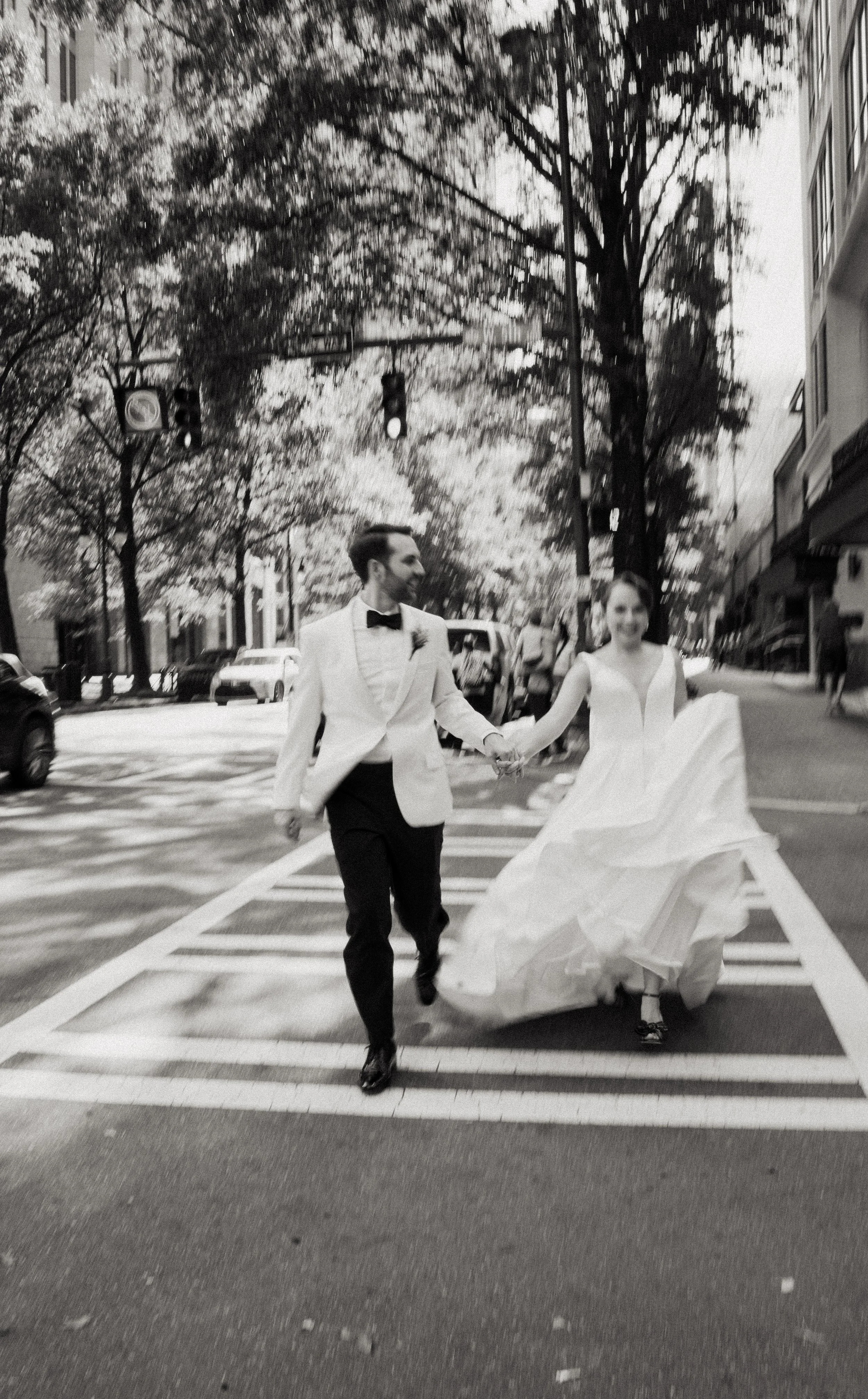 A newlywed couple, dressed in wedding attire, holding hands and walking across a city street at a crosswalk, with trees and buildings in the background.
