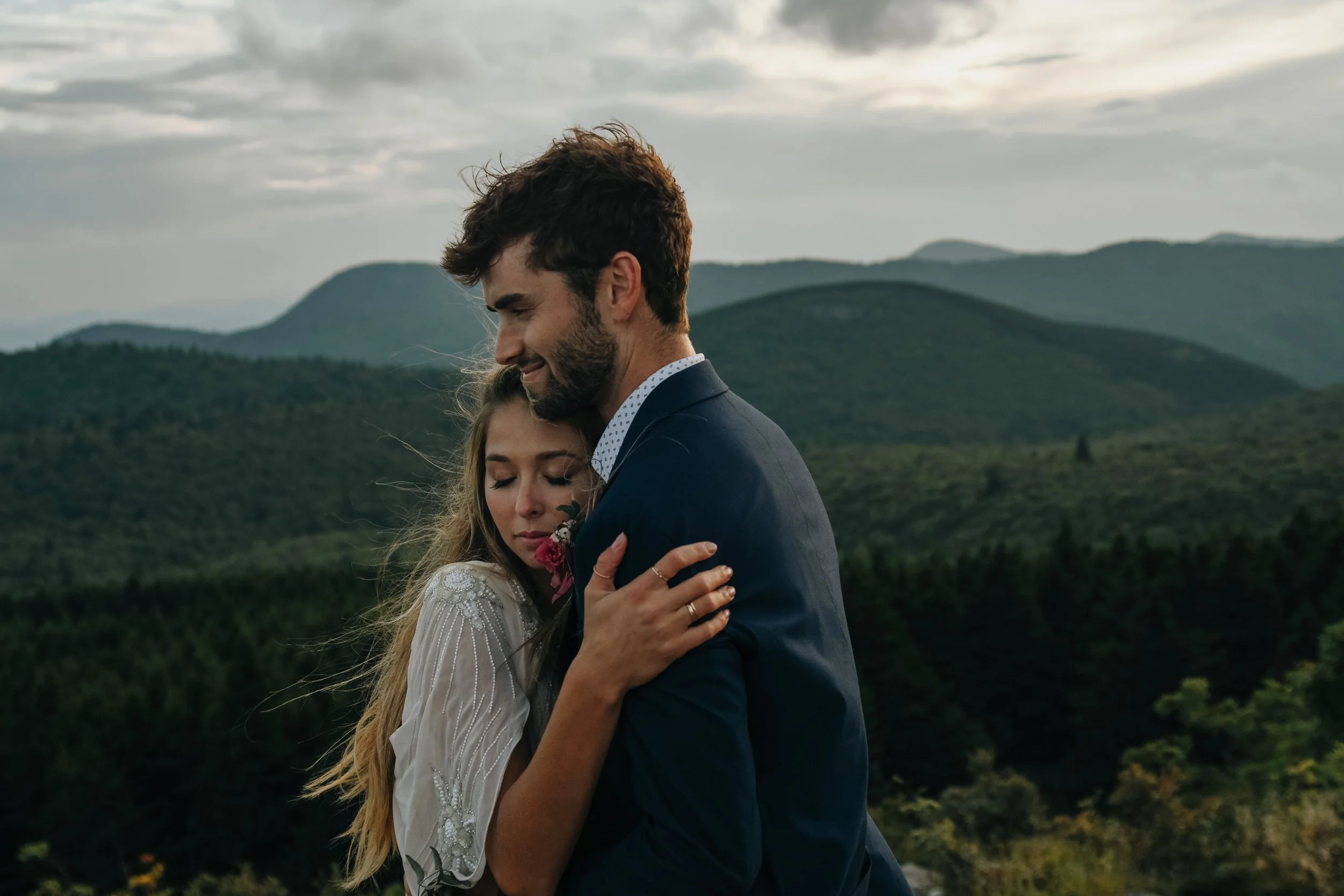 A young couple embraces outdoors in a scenic mountainous area during sunset, with mountains and cloudy sky in the background.