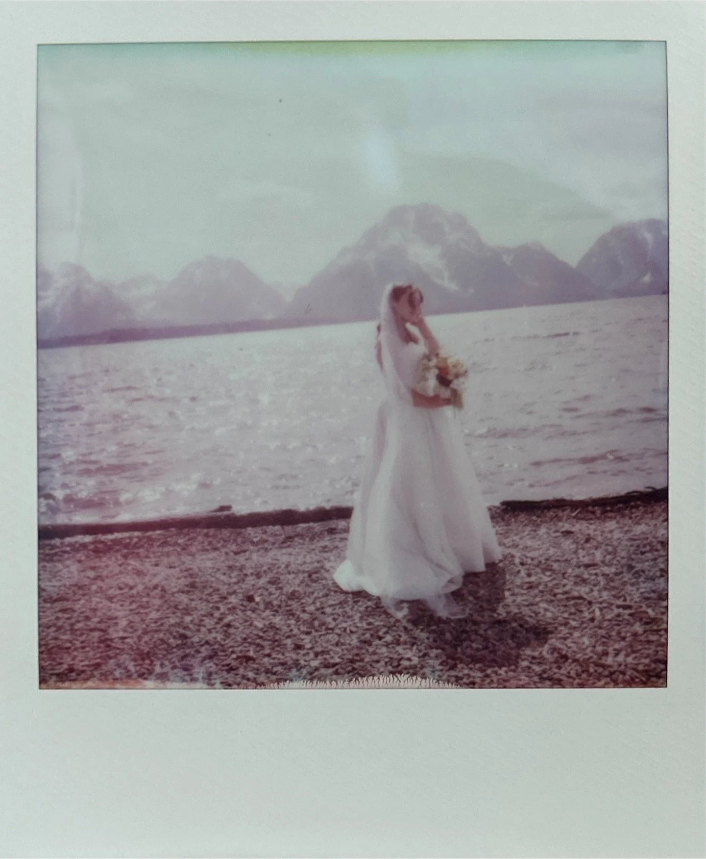 A woman in a white gown holding a bouquet stands on a rocky shore by a lake with mountains in the background.