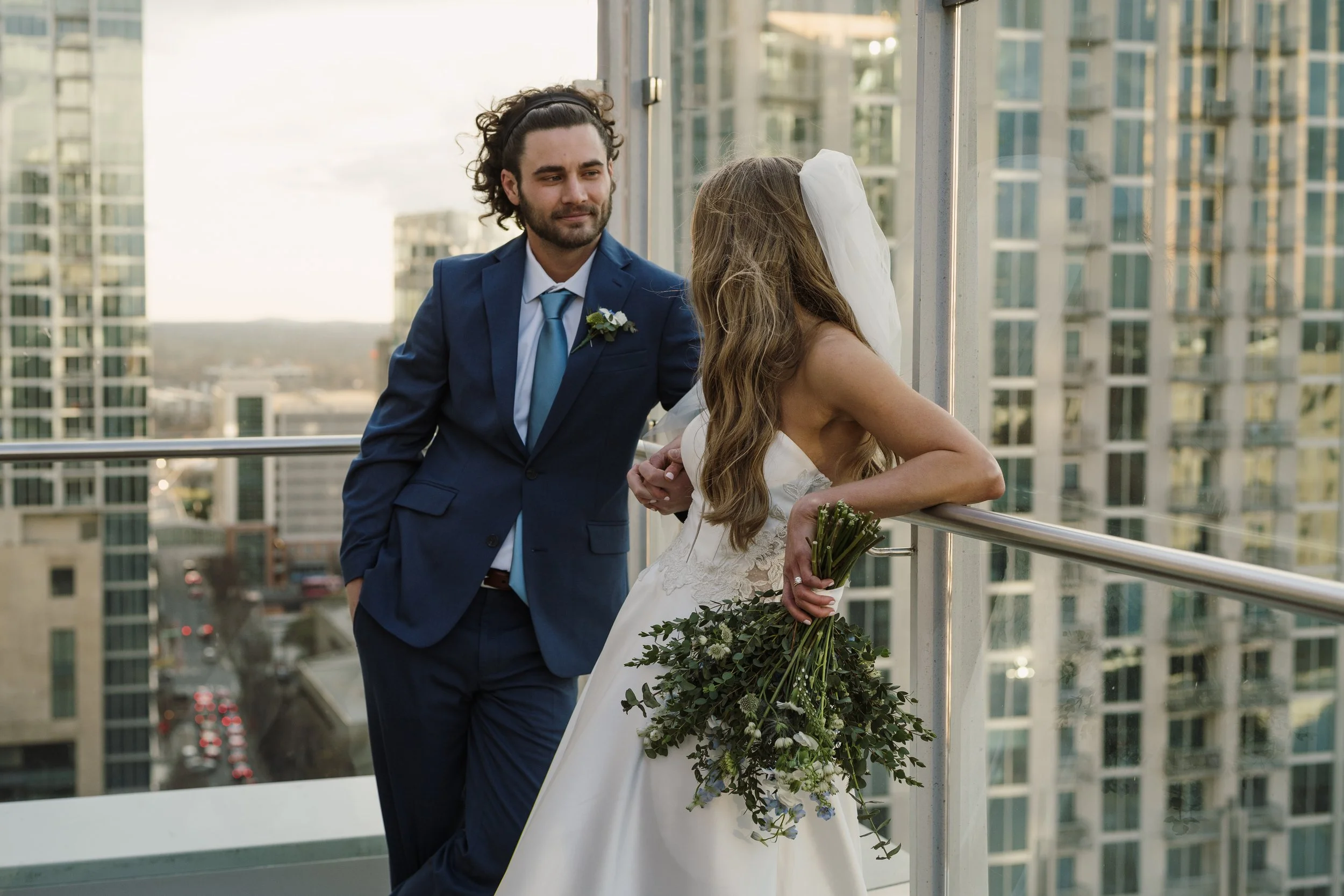 A bride and groom on a balcony with city buildings in the background. The groom is in a blue suit with a boutonniere, and the bride is wearing a white wedding dress, holding a bouquet of greenery.