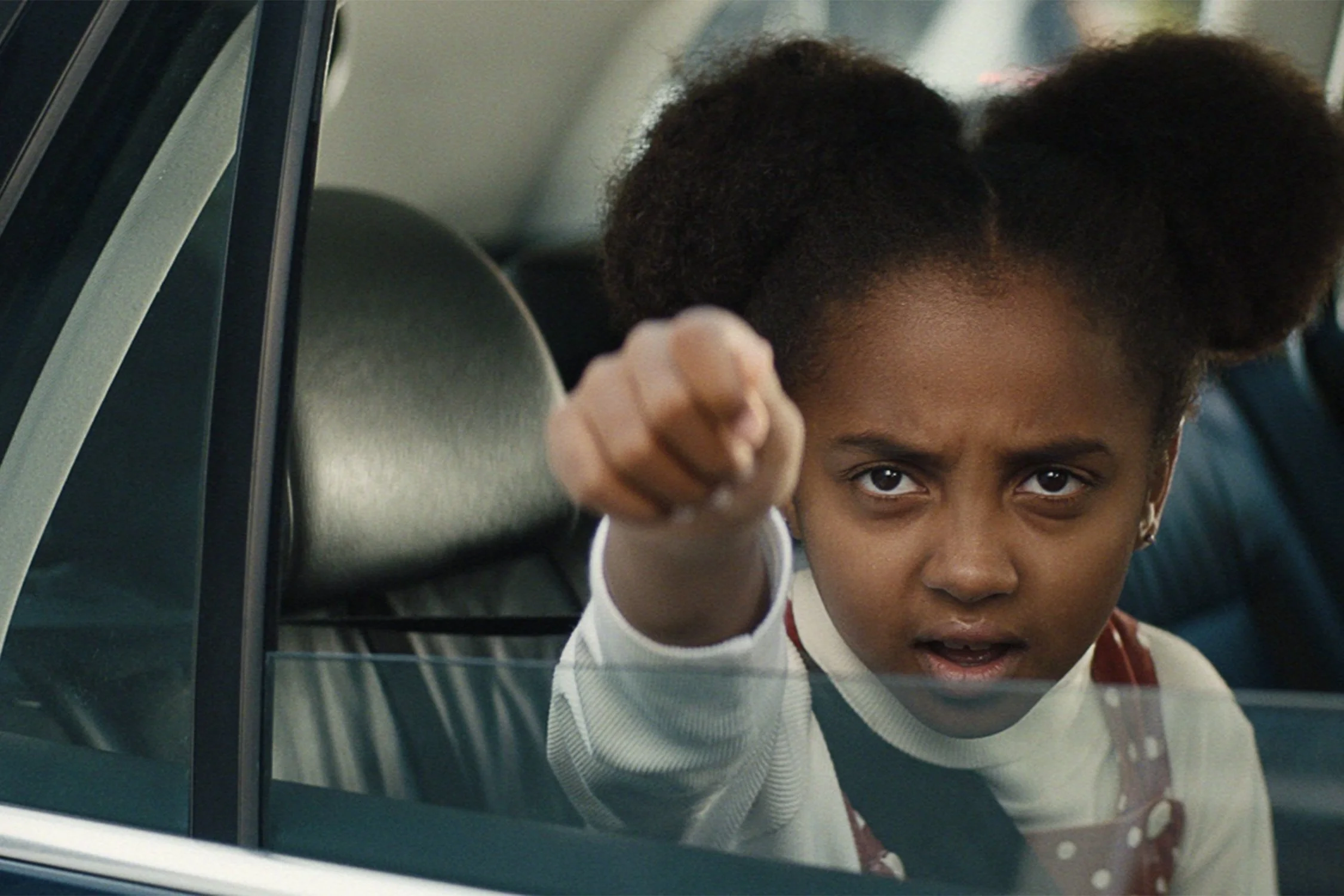 A young girl with puffed hair and a striped shirt points assertively through a car window.