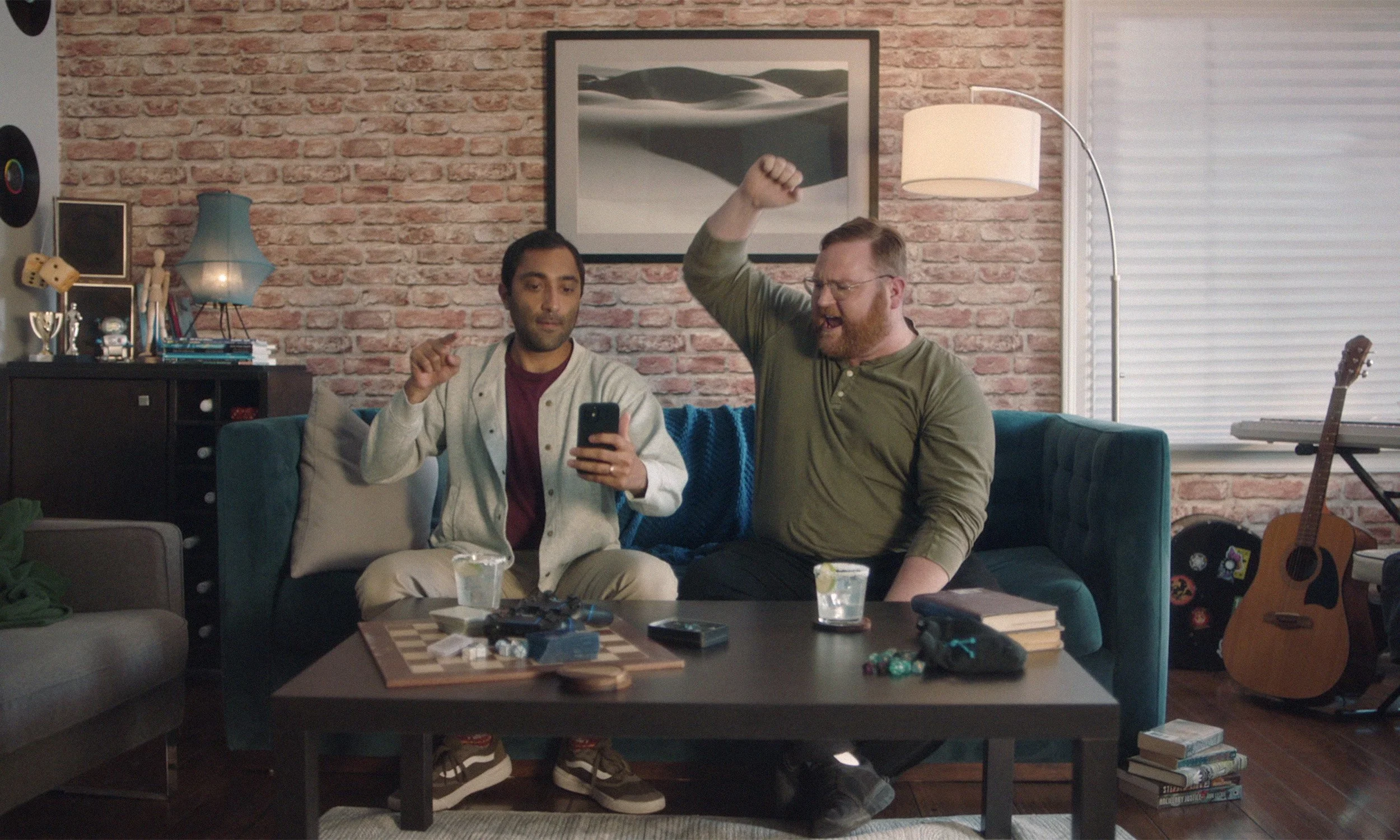 Two men sitting on a teal sofa, one is looking at a smartphone while the other is excitedly raising his arm. They are in a living room with a brick wall, guitar, and books around them.