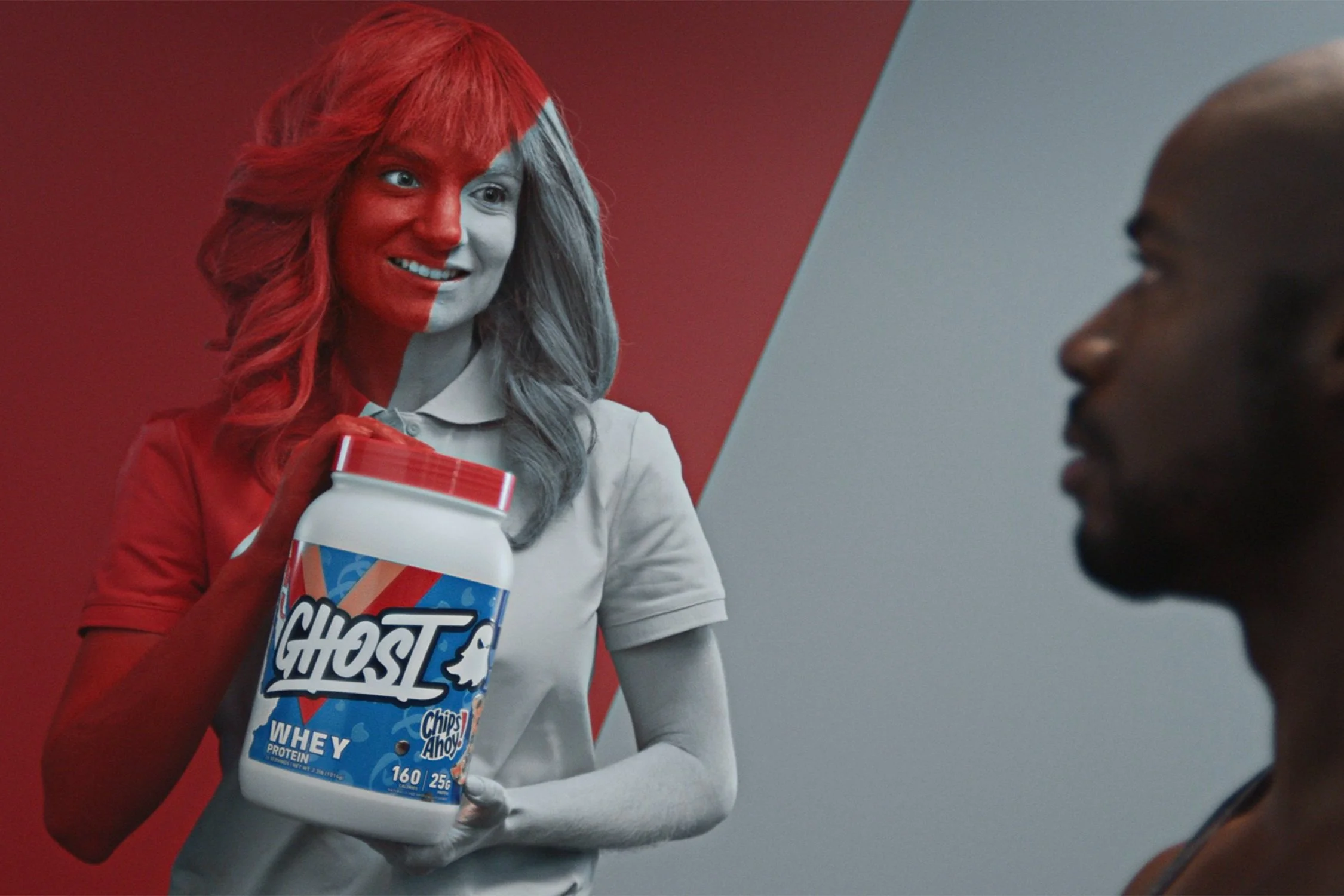 A woman with half of her face painted red and the other half gray, holding a container of Ghost whey protein powder, smiling and looking at a man in profile view against a red and gray background.