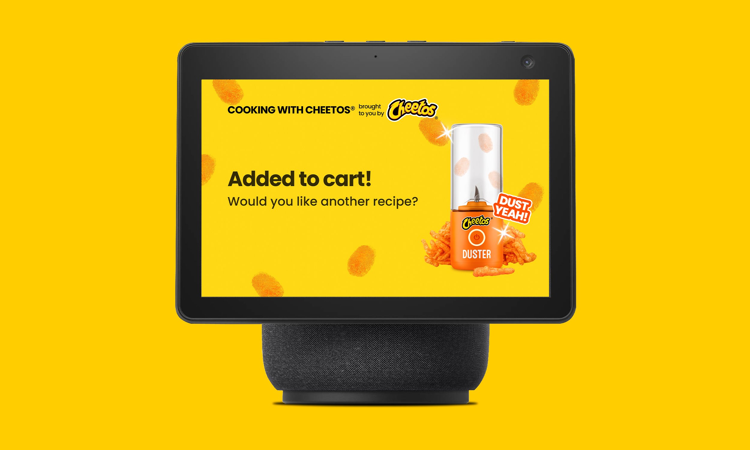 Digital display screen with yellow background showing a message 'Added to cart! Would you like another recipe?' next to a small orange kitchen appliance labeled 'Duster' with a Sparkle icon and a group of Cheetos snack pieces around it.